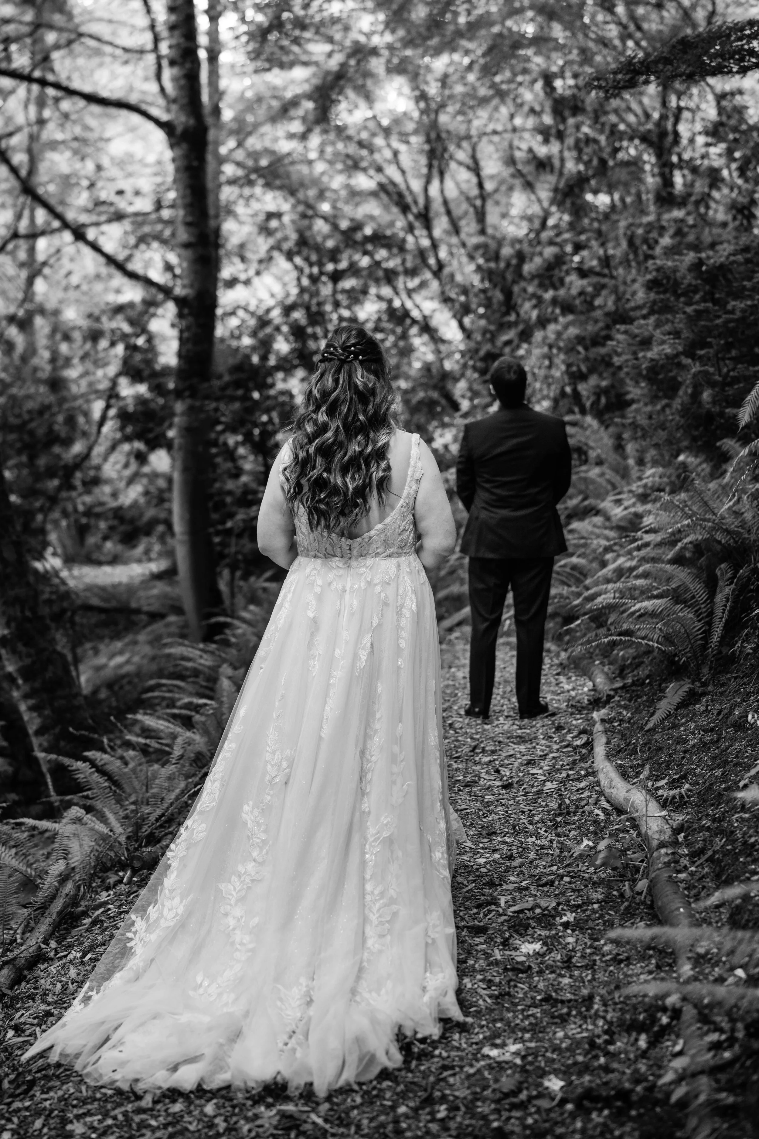 A black and white photo of a bride in a wedding dress and a groom in a suit, walking away through a wooded forest trail.