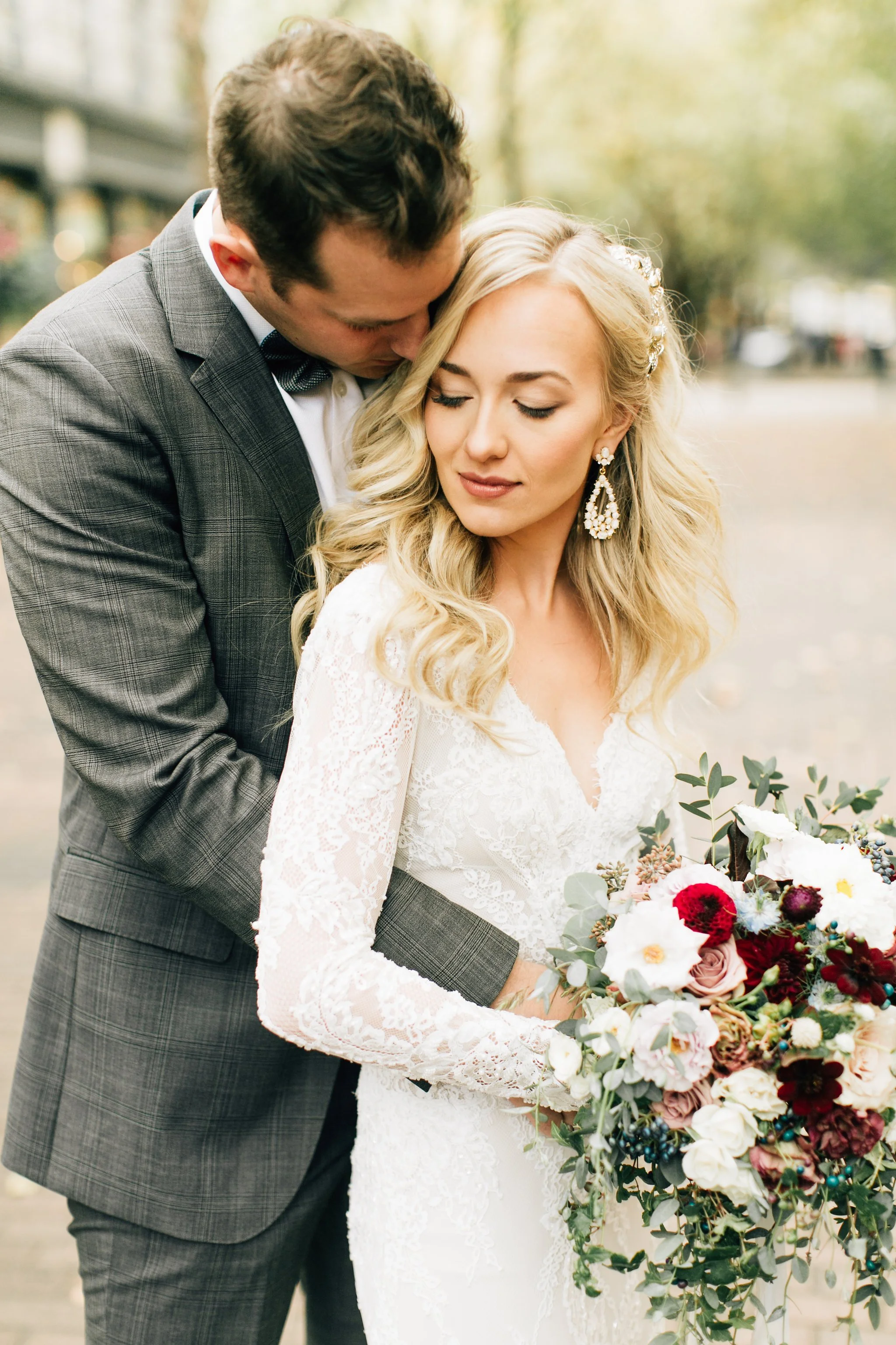 A bride and groom sharing a tender moment outdoors, with the groom kissing the bride's forehead, she has long wavy blonde hair with floral accessories, and is holding a large bouquet of flowers.
