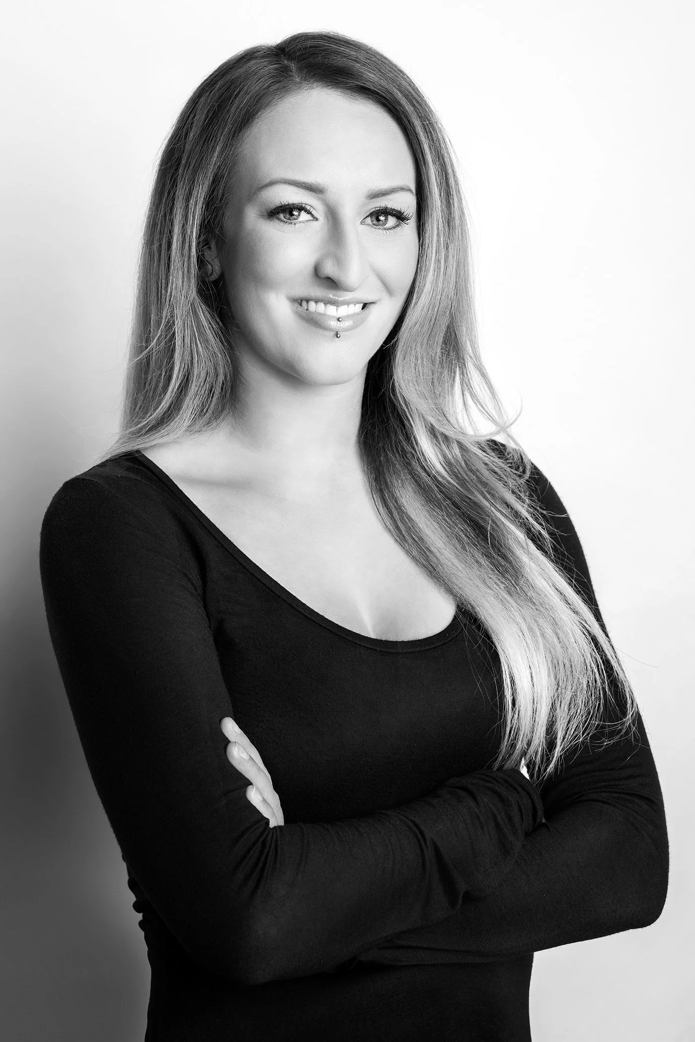 Black and white portrait of a woman with long hair, smiling, wearing a long-sleeved black top, and standing against a light background.