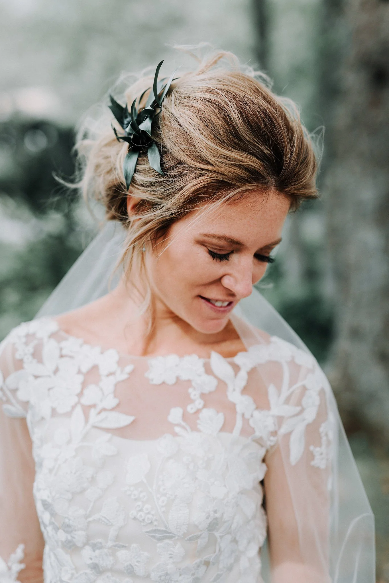 A bride with blonde hair styled in an updo, adorned with a black ribbon and greenery, wearing a white lace wedding dress with floral patterns and sheer long sleeves.