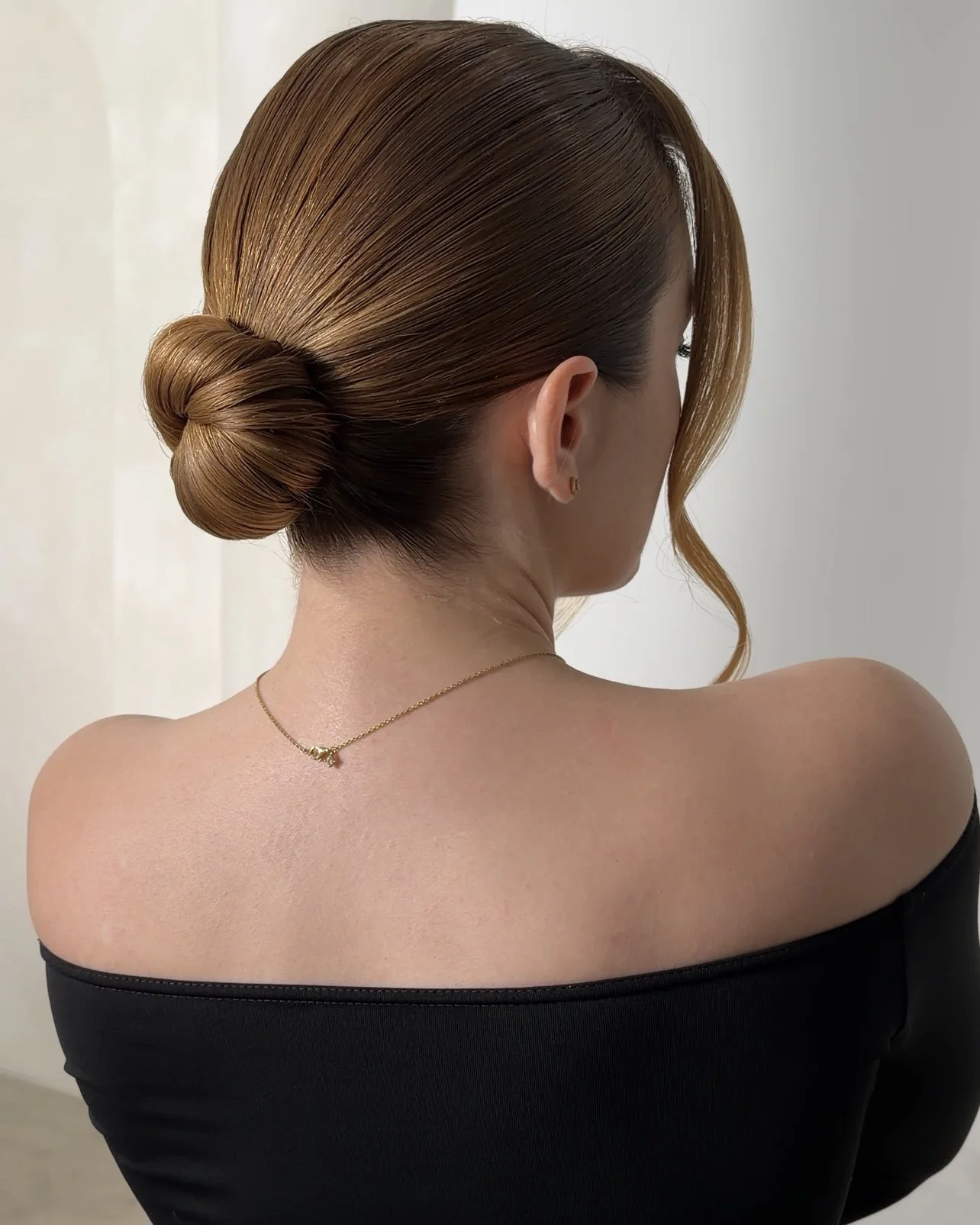 A woman with smooth, styled brown hair in a bun and a loose curl hanging near her face, wearing a black off-the-shoulder top and a delicate gold necklace, with a plain light-colored background.