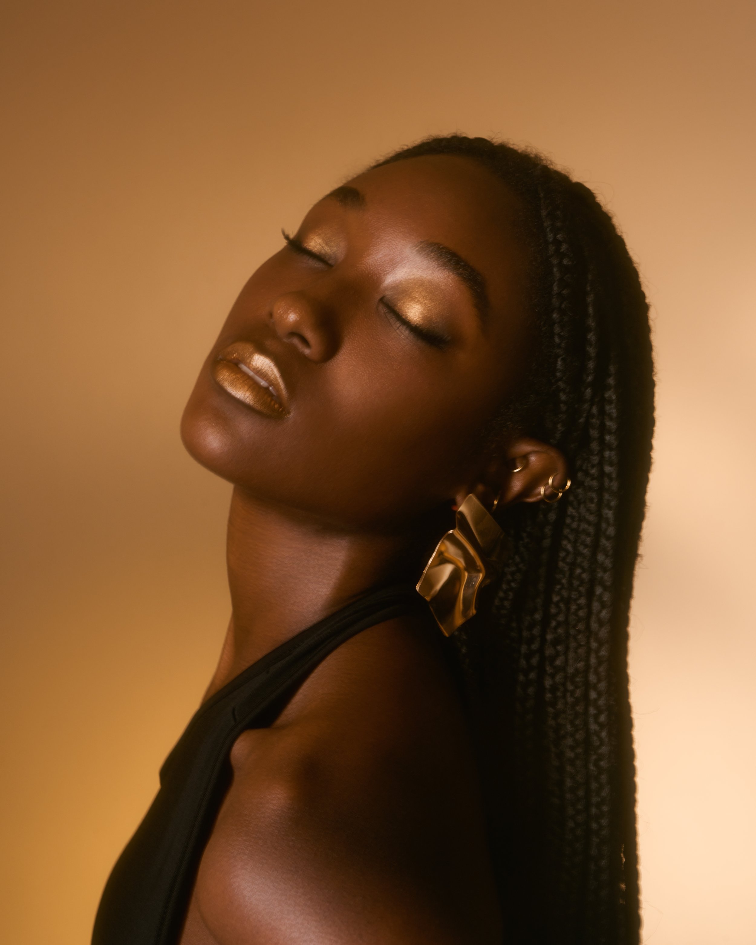 A close-up of a woman with dark skin, closed eyes, gold eye makeup, and gold lipstick, wearing a large gold earring, with her head tilted back against a beige background.