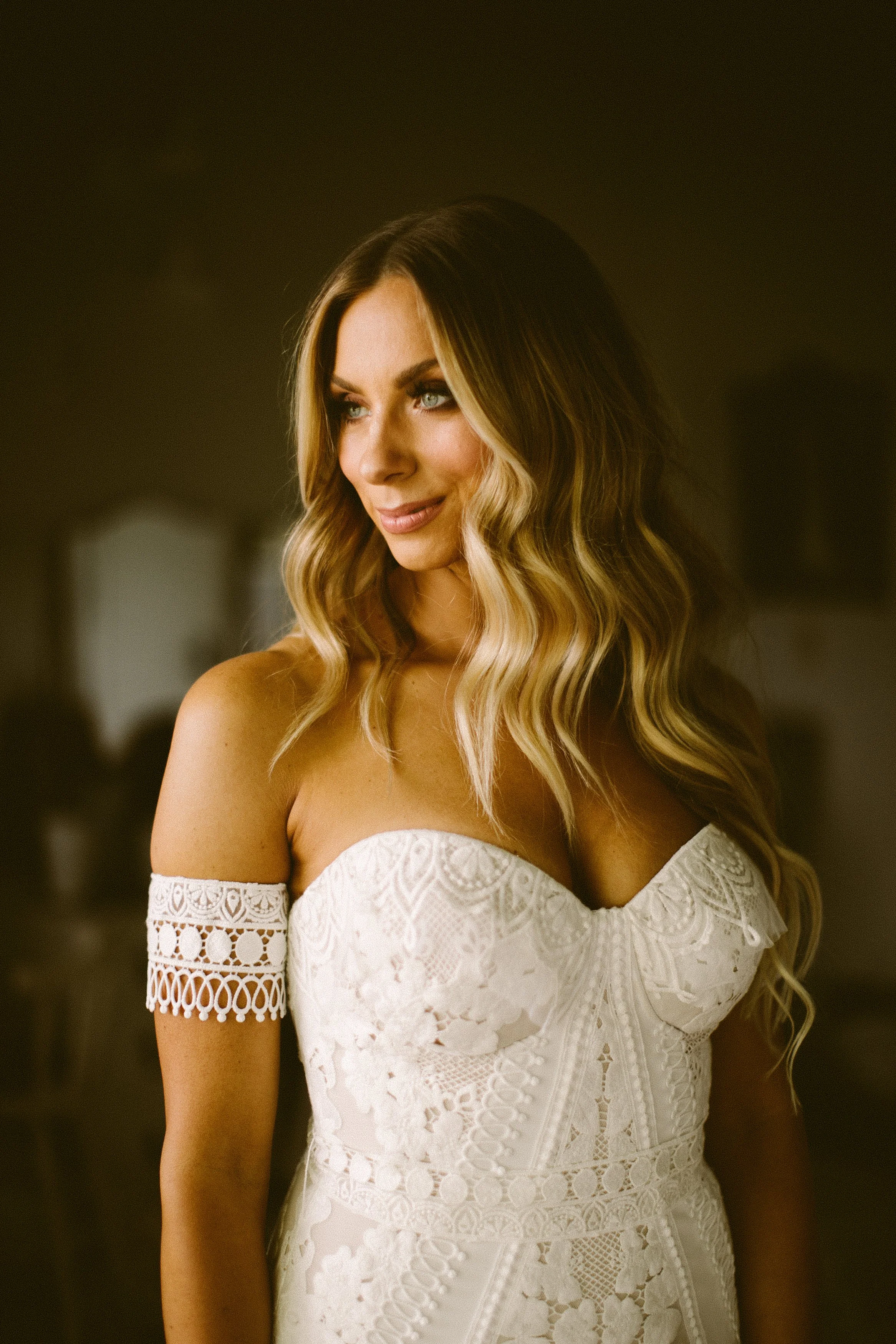 A woman with long wavy hair, wearing a white lace off-the-shoulder dress, smiling softly, in a dimly lit indoor setting.