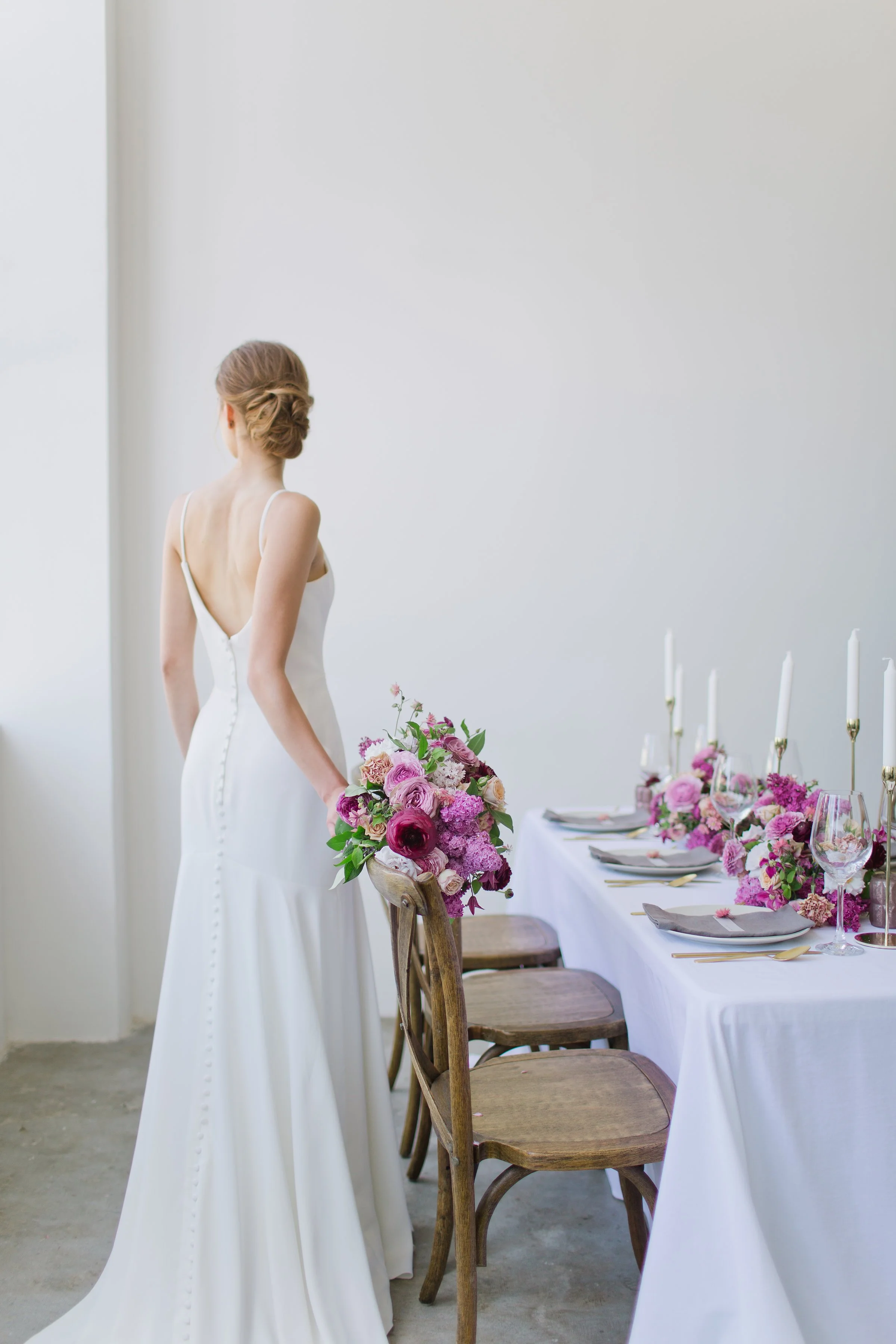 A woman in a white wedding dress holding a bouquet of pink and purple flowers, standing in a decorated dining area with a table set for a special occasion.