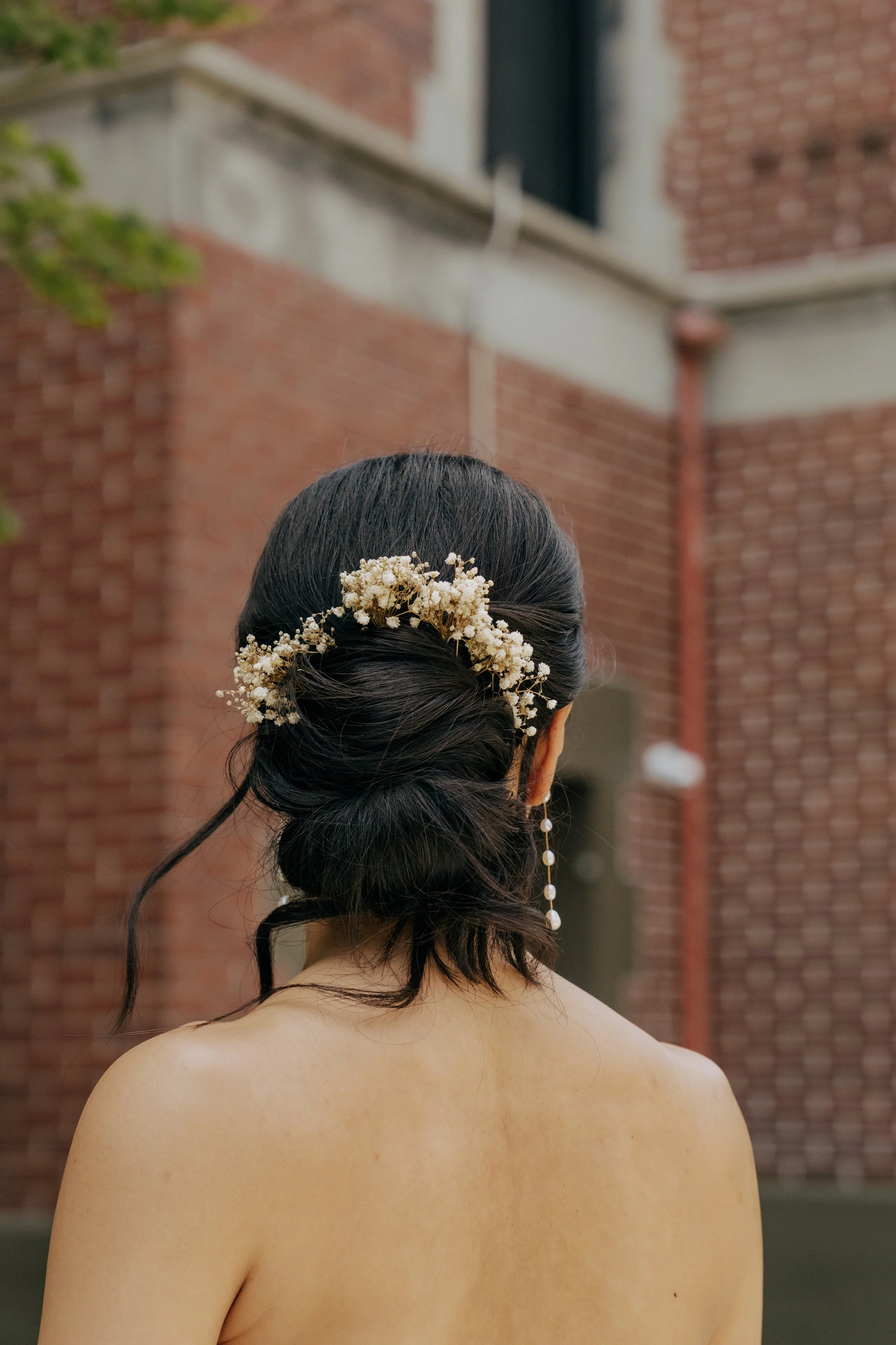 A woman with dark hair styled in an elegant updo adorned with white baby's breath flowers and pearl earrings, facing away outdoors with a brick building in the background.