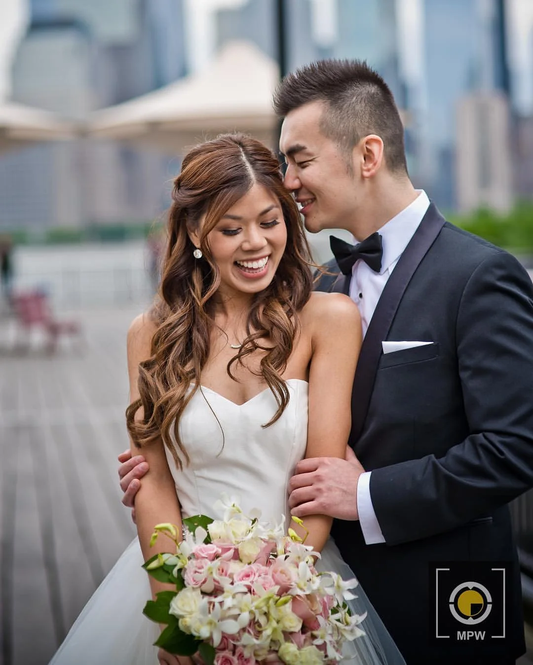 A newlywed couple in wedding attire, smiling with eyes closed, standing outdoors in an urban setting, with the woman holding a bouquet of pink and white flowers.