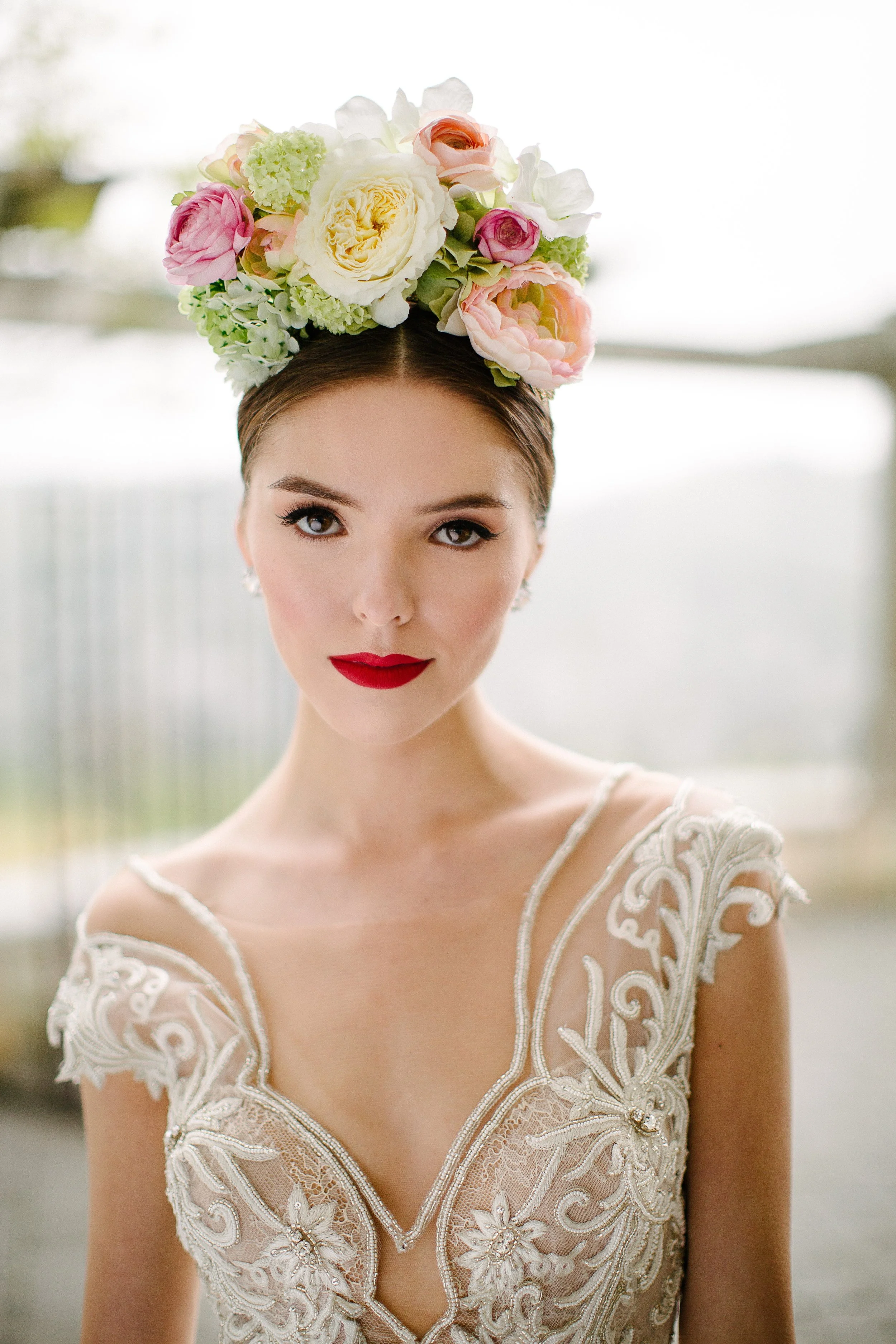 Woman wearing a floral crown made of white, pink, and peach flowers, dressed in a lace wedding gown with intricate embroidery and beadwork.
