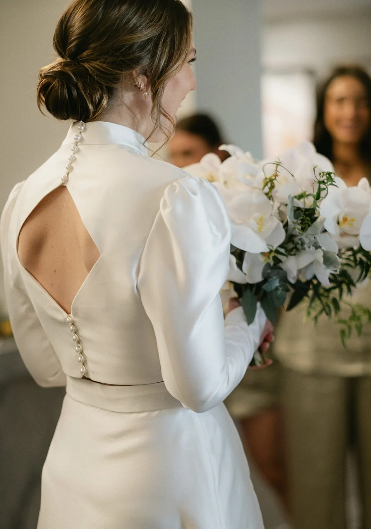 A woman in a white dress holding a bouquet of white orchids, with several women in the background, at a wedding or formal event.