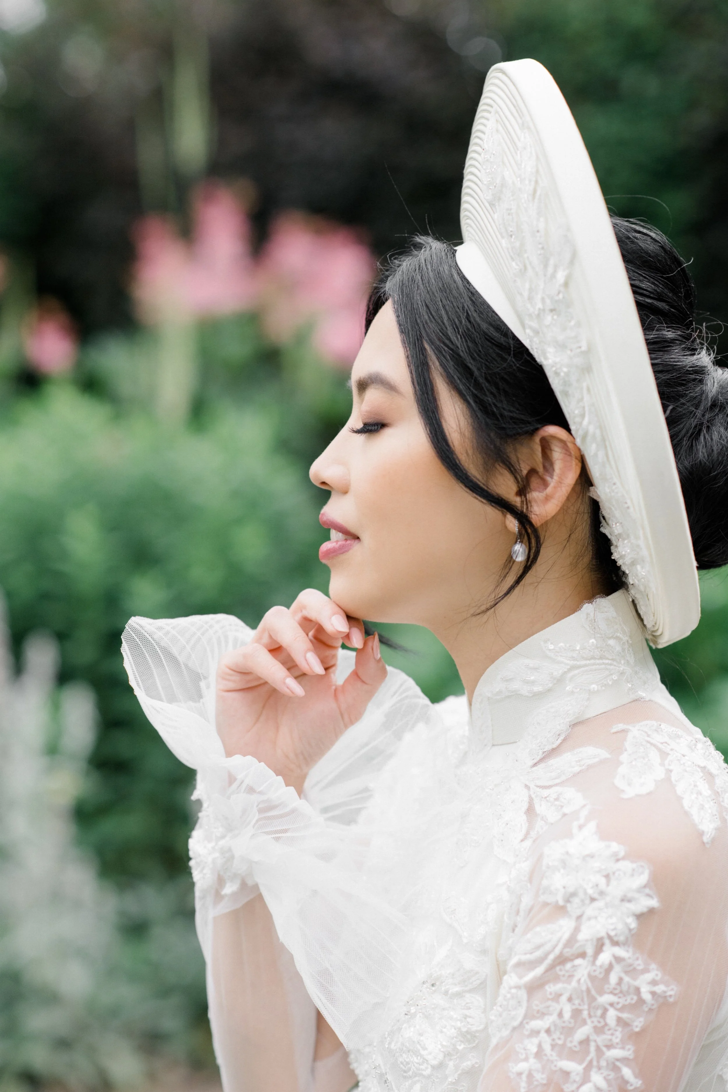 A woman in a white lace dress with a wide-brim hat, standing outdoors with a green background and pink flowers in the blurred distance.