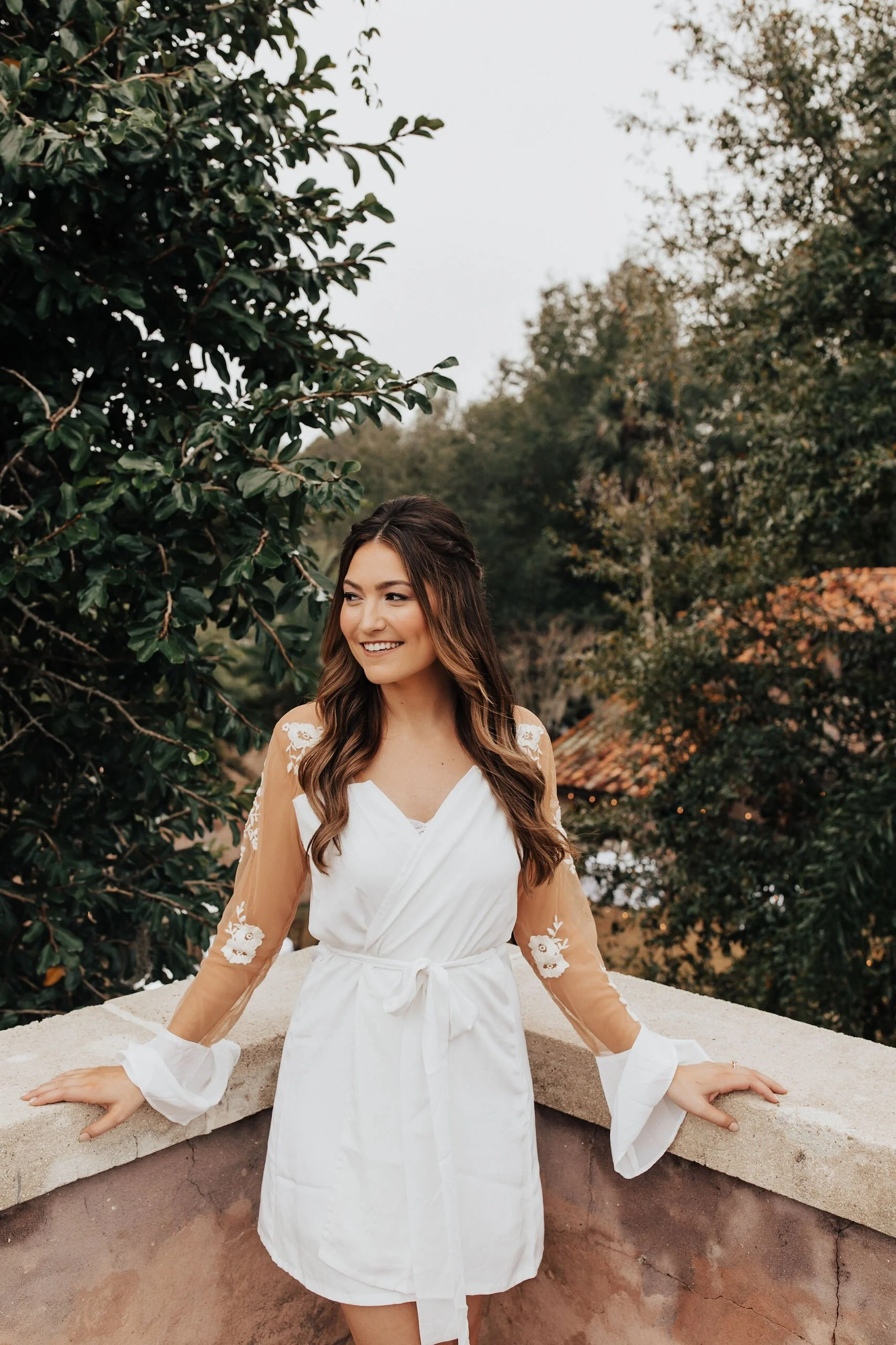 Young woman in a white dress with sheer, embroidered sleeves, smiling while leaning on a stone ledge outdoors surrounded by trees.