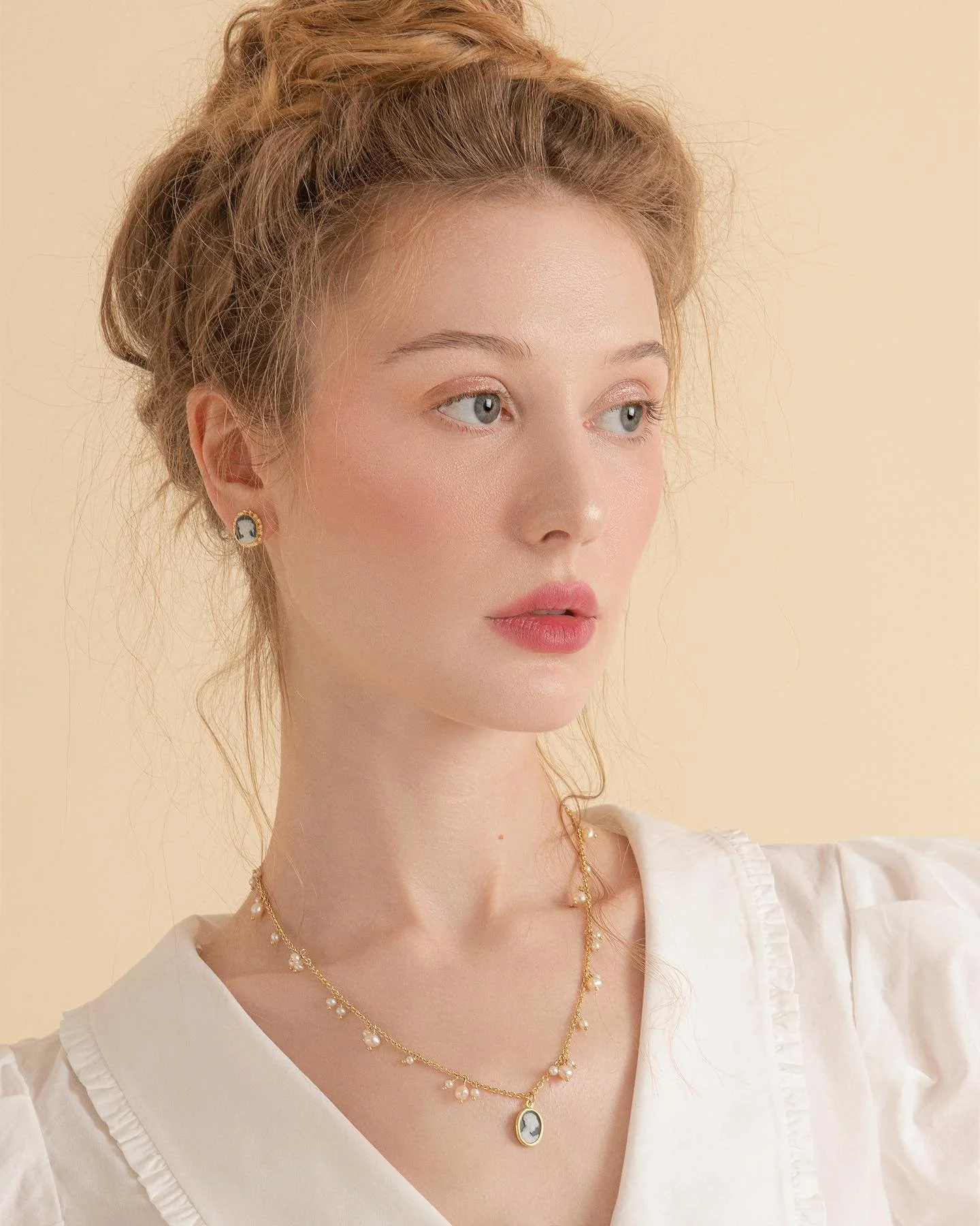 Portrait of a young woman with red hair styled in a messy updo, wearing pearl earrings and a gold layered necklace, dressed in a white top, against a neutral beige background.