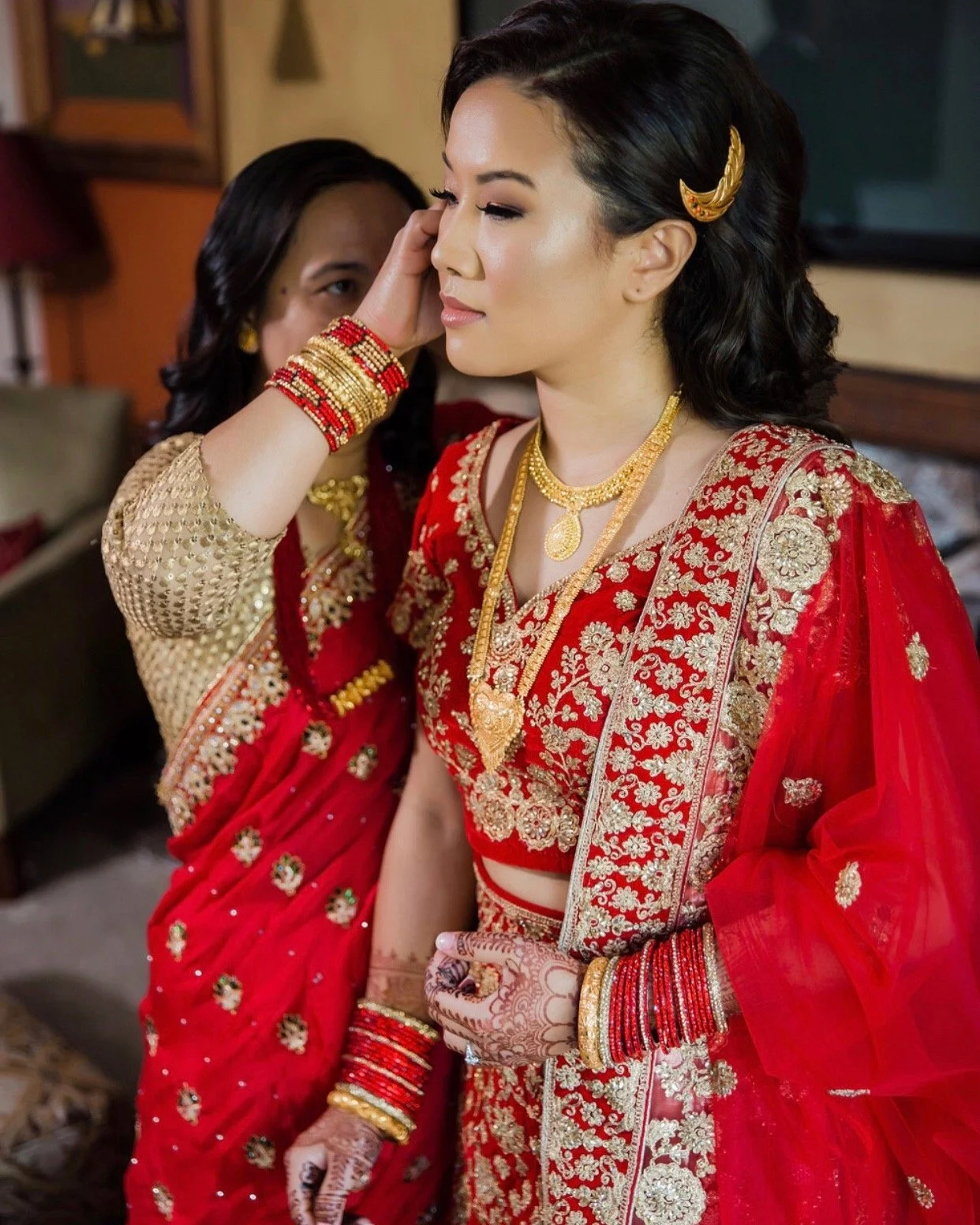 Two women dressed in traditional Indian wedding attire, with one woman applying jewelry to the other.