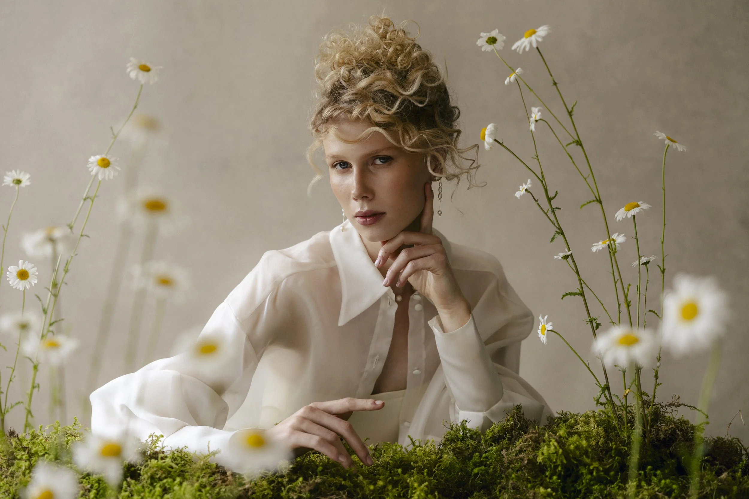 A woman with blonde curly hair styled in an updo, wearing a white silk blouse, surrounded by daisies and moss, posing thoughtfully with her hand near her face.