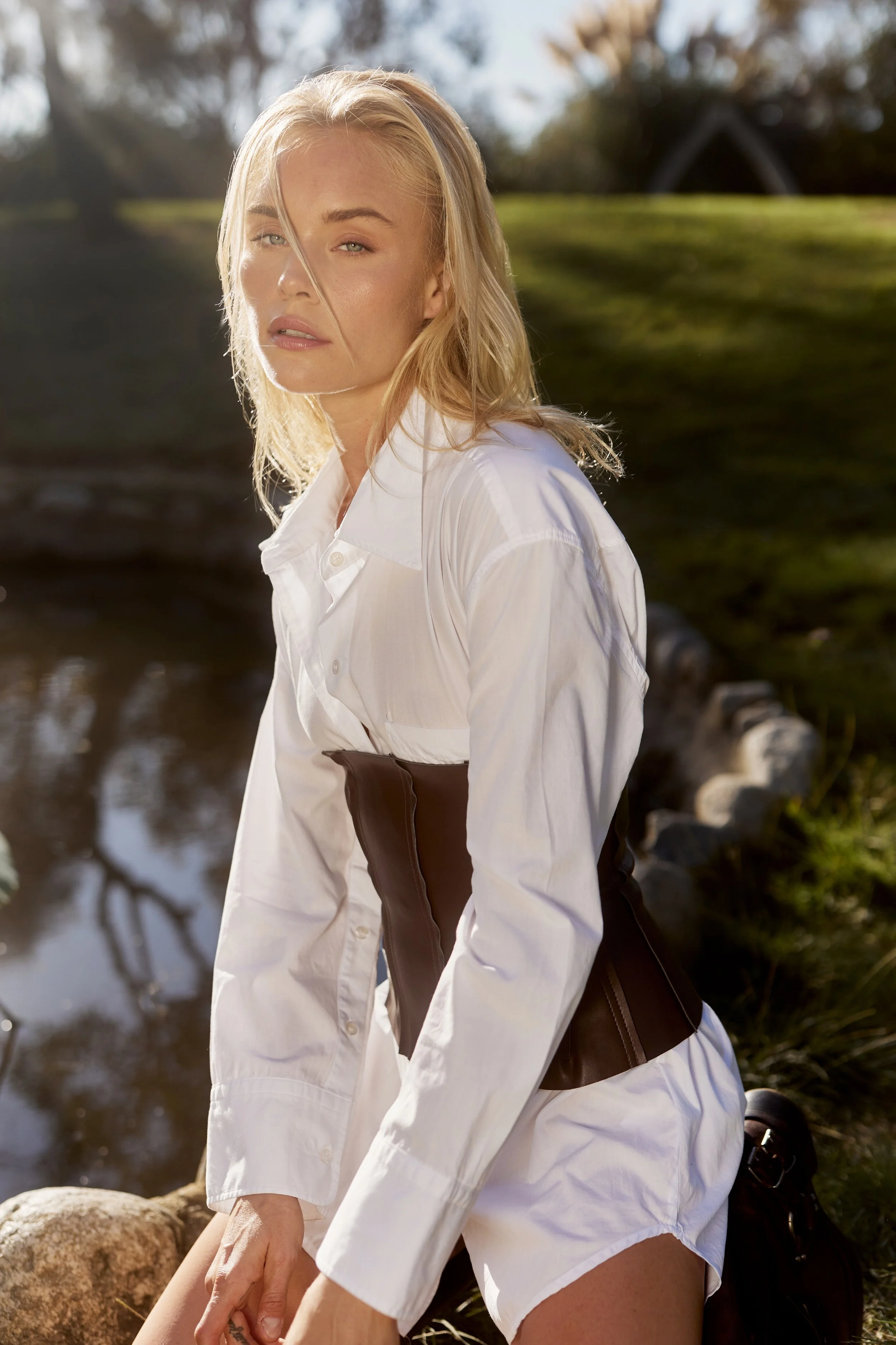 A woman with blonde hair wearing a white shirt and brown pants, sitting outdoors near a body of water with rocks and trees in the background, bathed in warm sunlight.