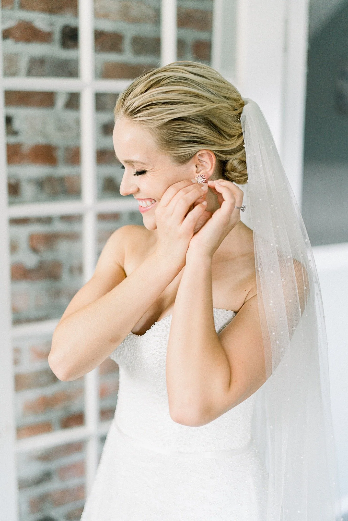 A bride with blonde hair styled in an elegant updo, wearing a white wedding dress and veil, smiling while putting on an earring in front of a brick wall and window.