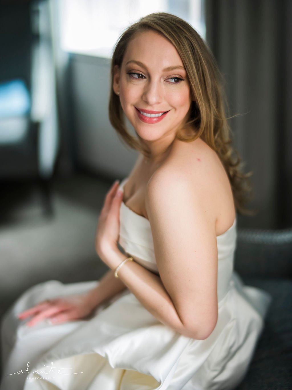 A woman with red hair sitting on a bed, smiling and looking over her shoulder, wearing a white strapless dress.
