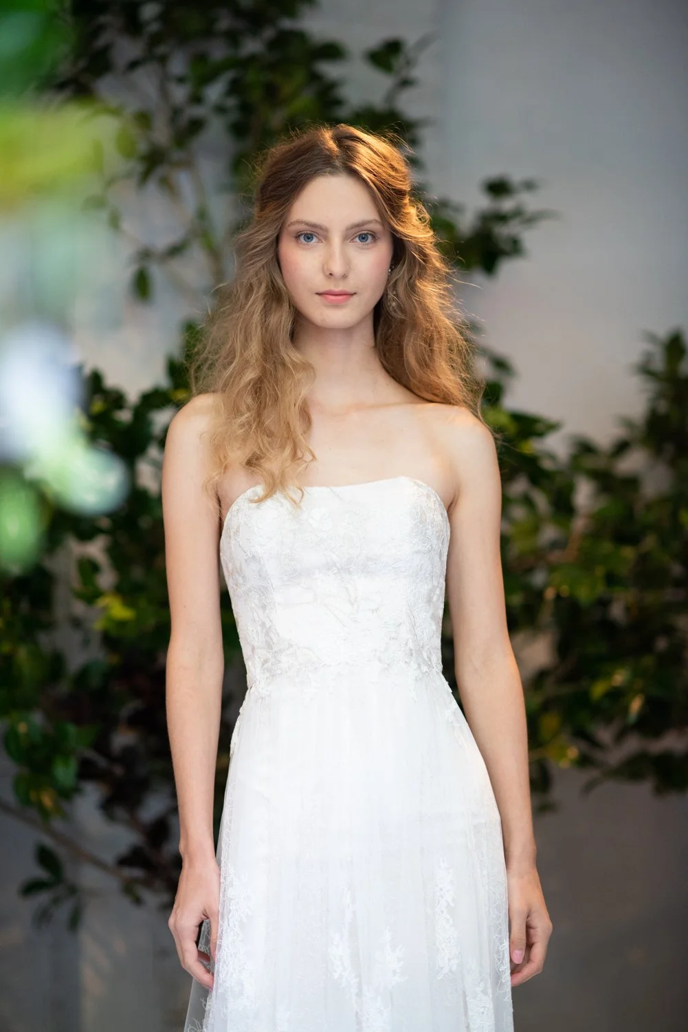 A young woman with long, wavy blonde hair, wearing a strapless white dress with lace details, standing outdoors with green foliage in the background.