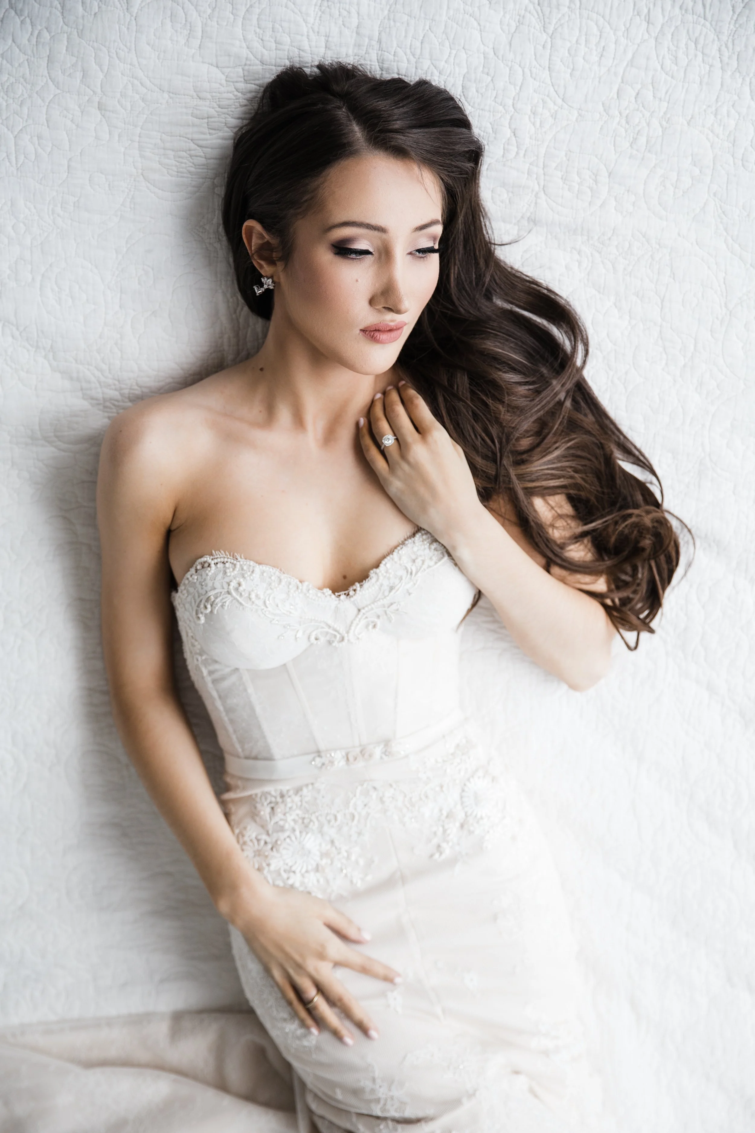 A woman in a white wedding dress lying on a quilted surface with long, dark hair, pearl earrings, and a ring on her left hand.