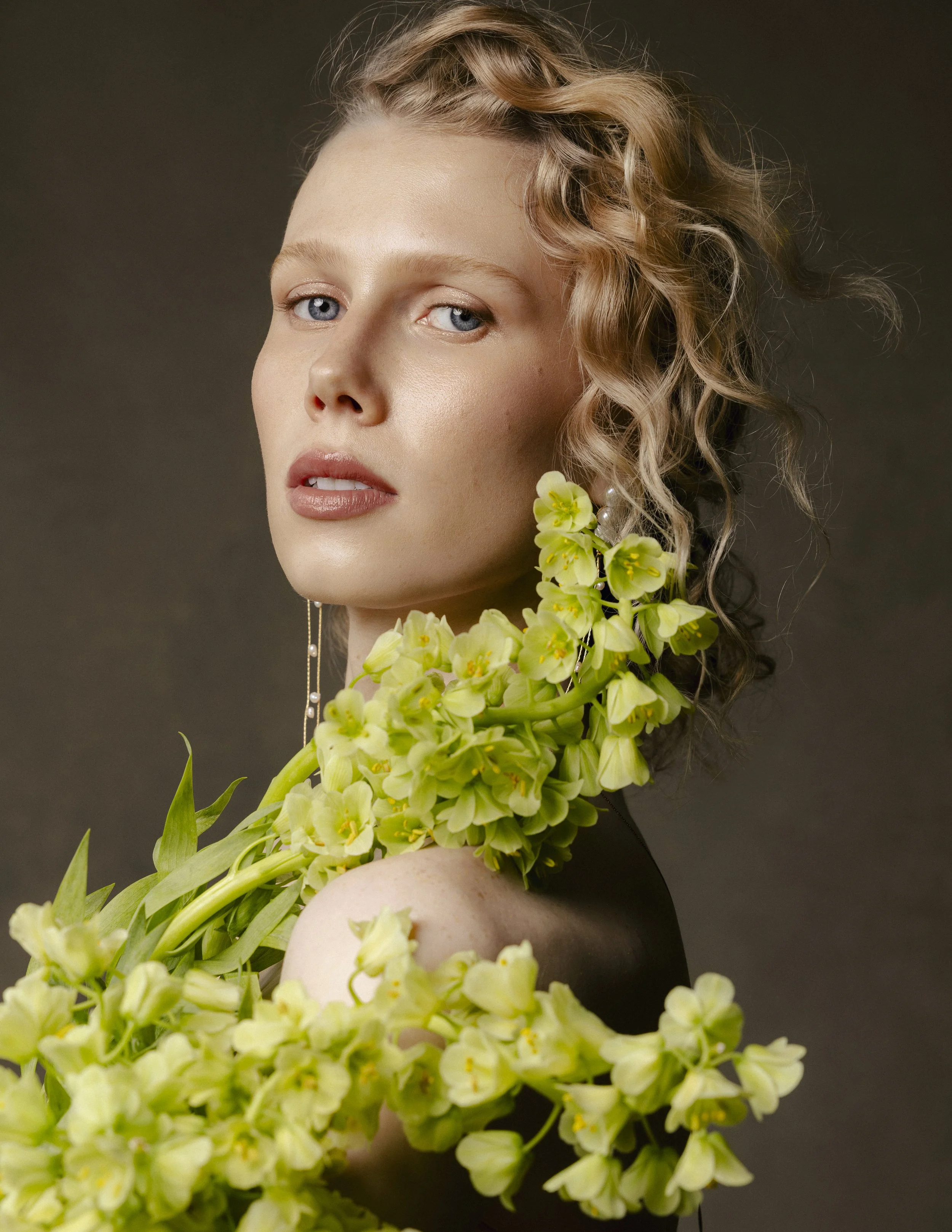 A woman with curly blonde hair and blue eyes, wearing makeup, earrings, and surrounded by yellow flowers, against a dark background.