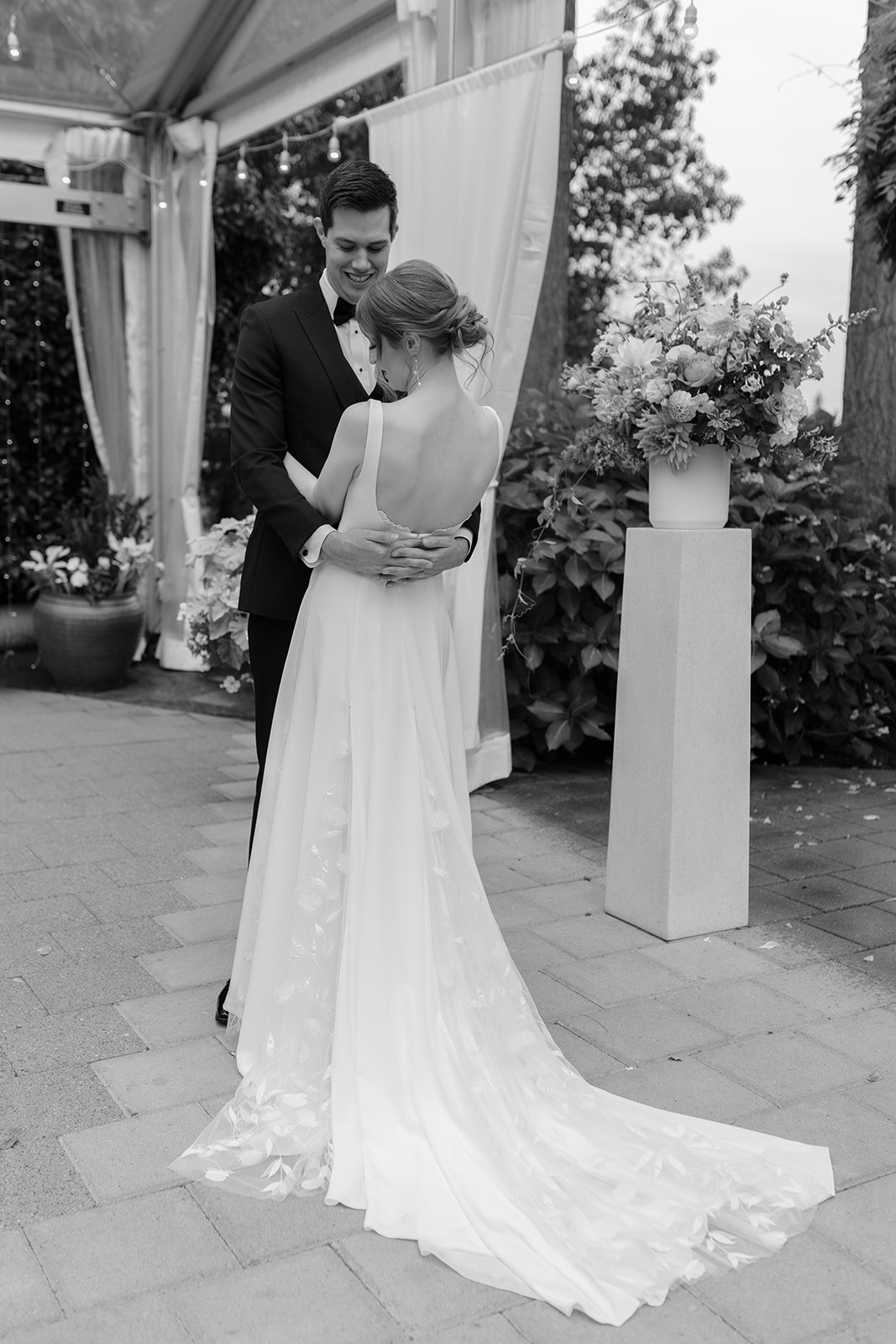 Black and white photo of a bride and groom at their wedding, dancing and smiling, outdoors with floral arrangements and a decorative backdrop.