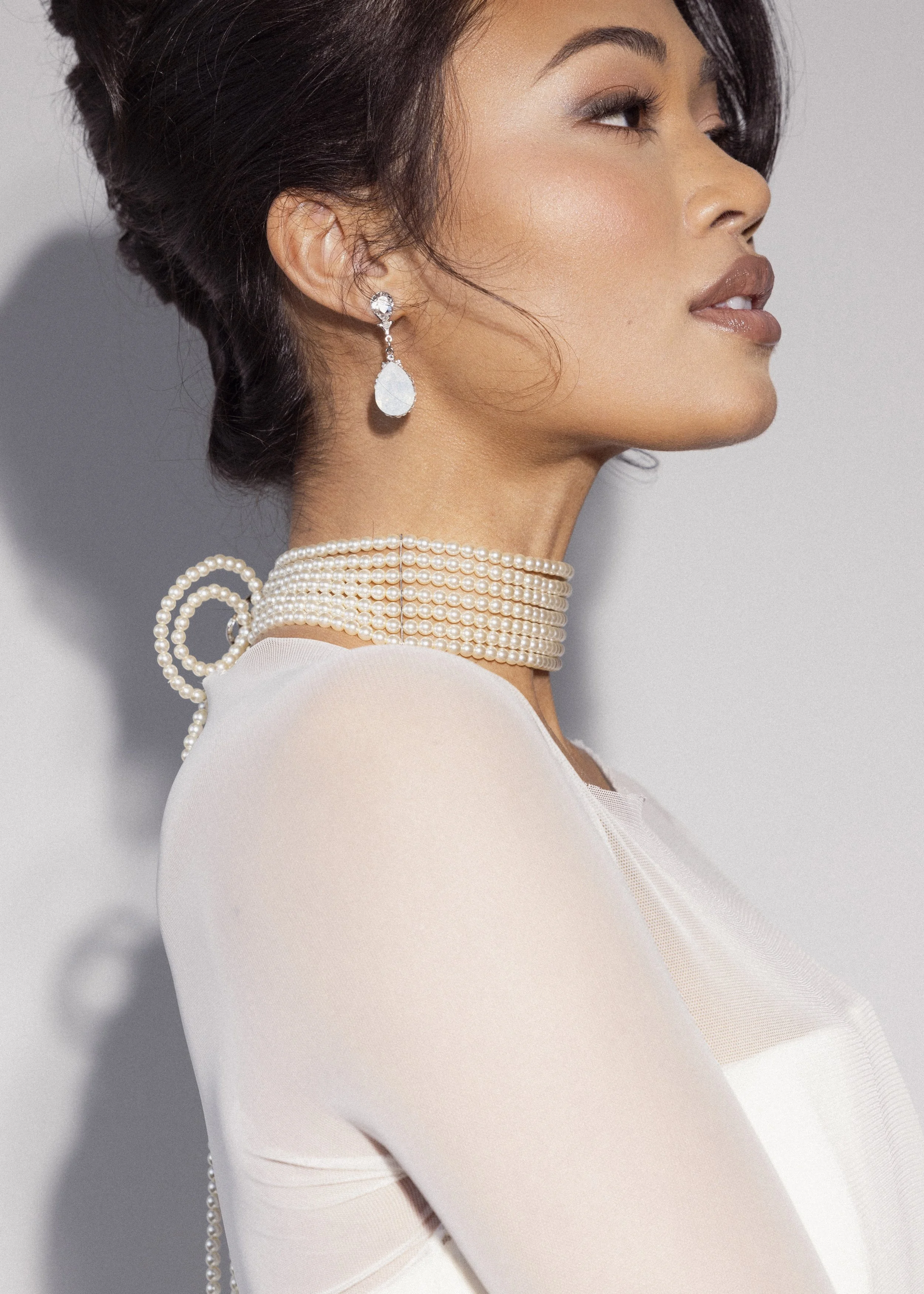 Side profile of a woman with short dark hair wearing a pearl necklace, pearl earrings, and a sheer white top.