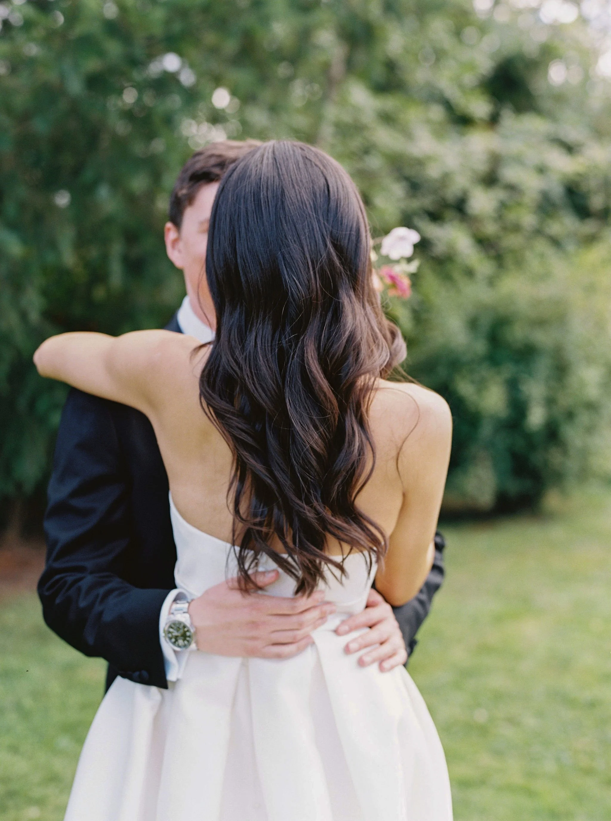 A couple in white wedding attire embracing outdoors with a green, blurred background.