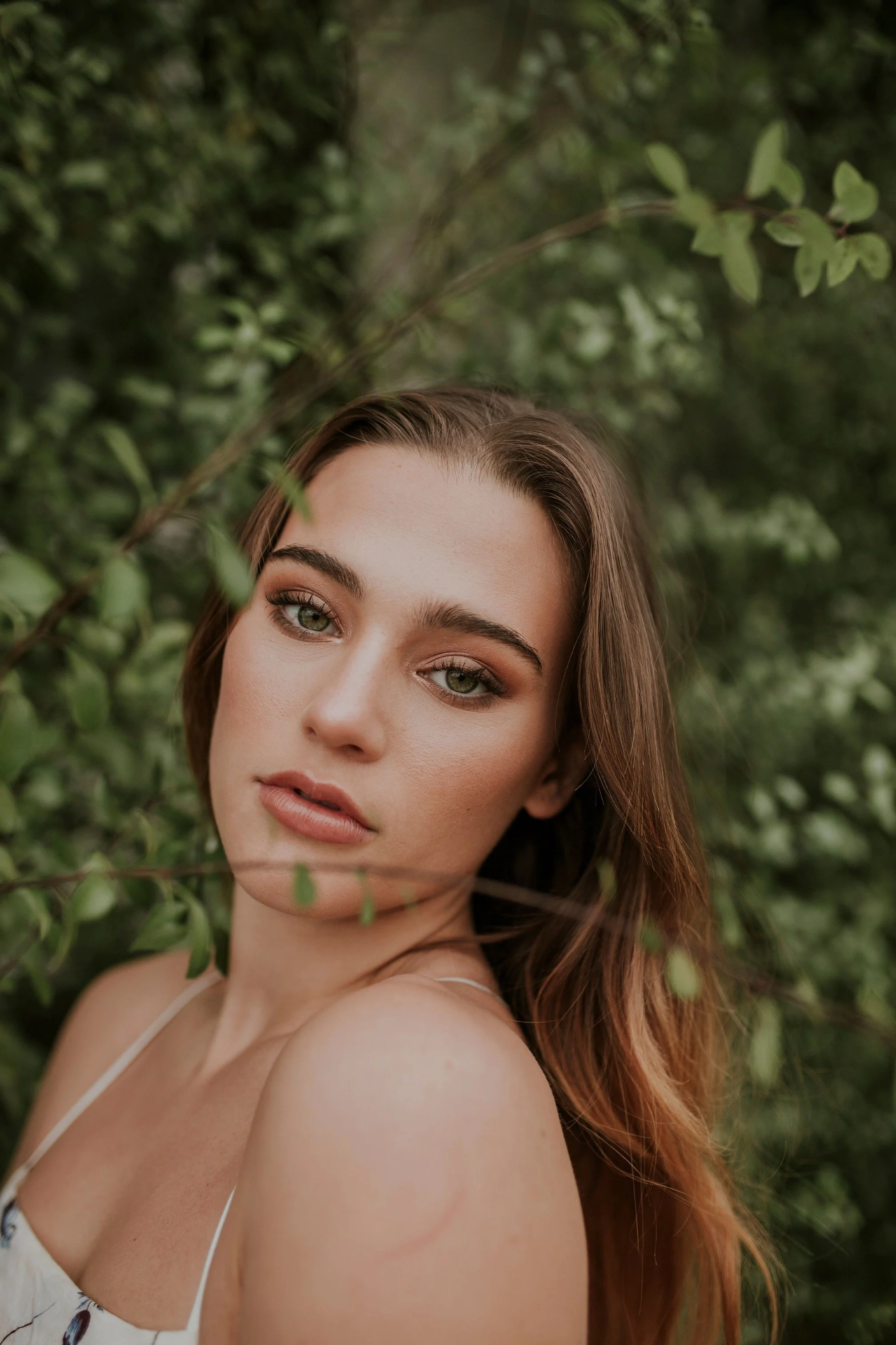 A young woman with long, wavy brown hair and light skin, gazing into the camera, surrounded by green foliage.