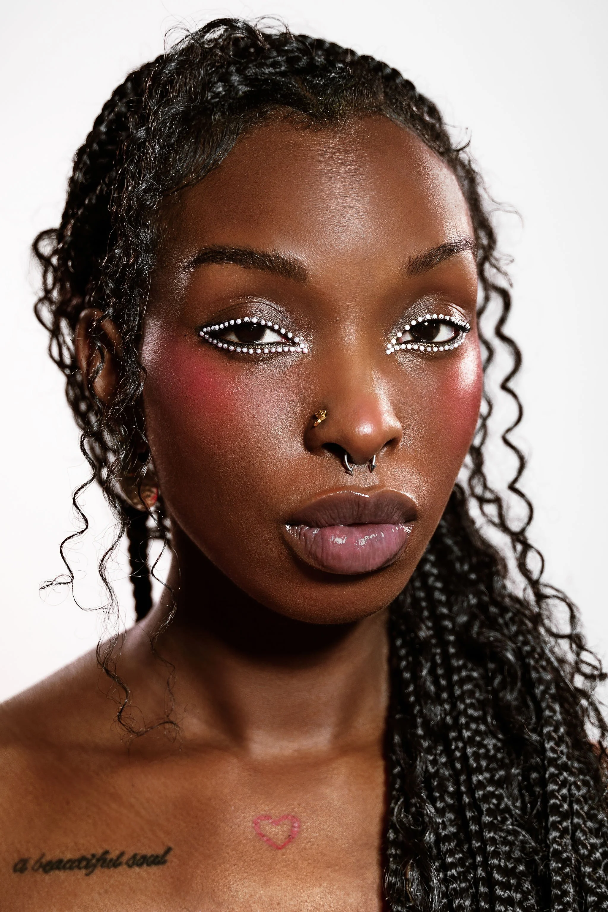 Close-up of a woman with curly black hair, wearing makeup with rhinestone accents around her eyes, a nose ring, and a chest tattoo, against a plain white background.