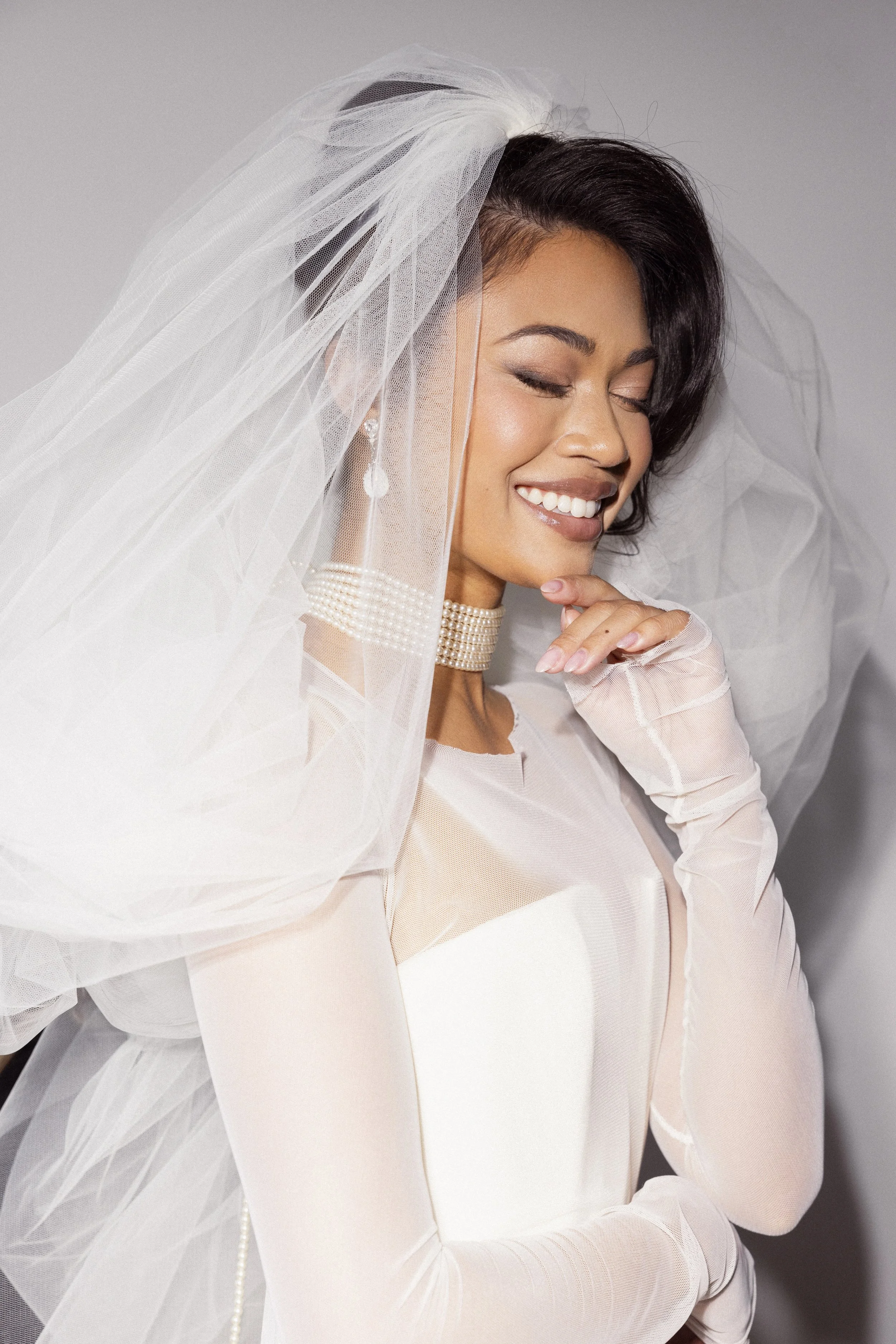 A smiling bride in a wedding dress, veil, and pearl jewelry, with her eyes closed and resting her right hand on her chin.
Makeup Katya
Hair Meghann
Photographer Andrew Parsons
