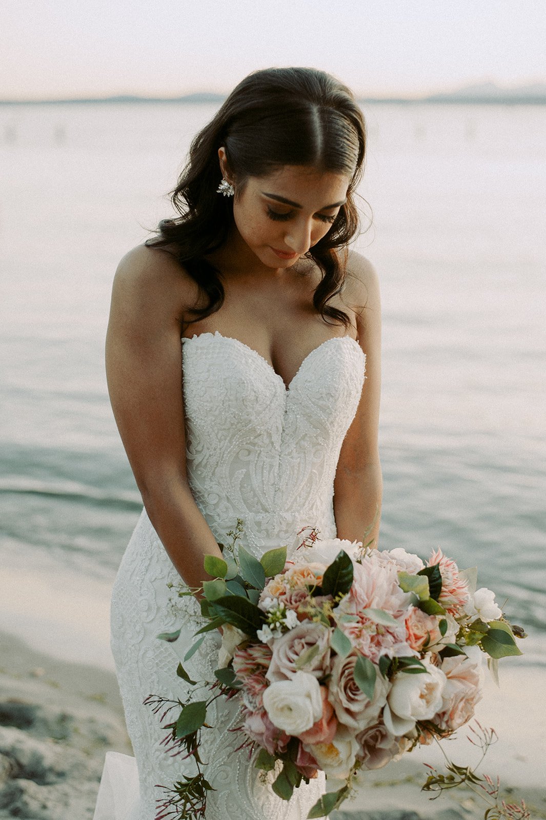 Bride in a white lace wedding gown holding a large bouquet of pink and white flowers with greenery, standing by a body of water during sunset.