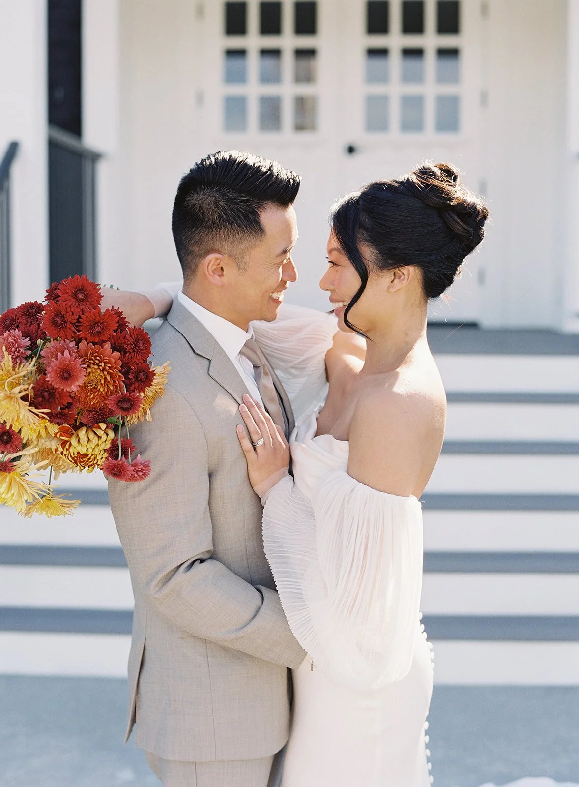 A wedding couple smiling and looking into each other's eyes; the groom is holding a bouquet of red and orange flowers, and they are standing outdoors on a staircase in front of a white building.