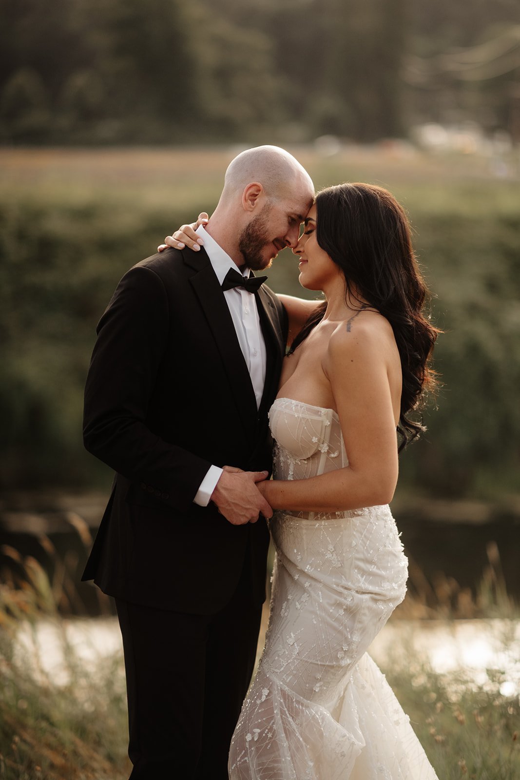 A bride and groom standing close together, touching foreheads and holding hands outdoors during sunset.