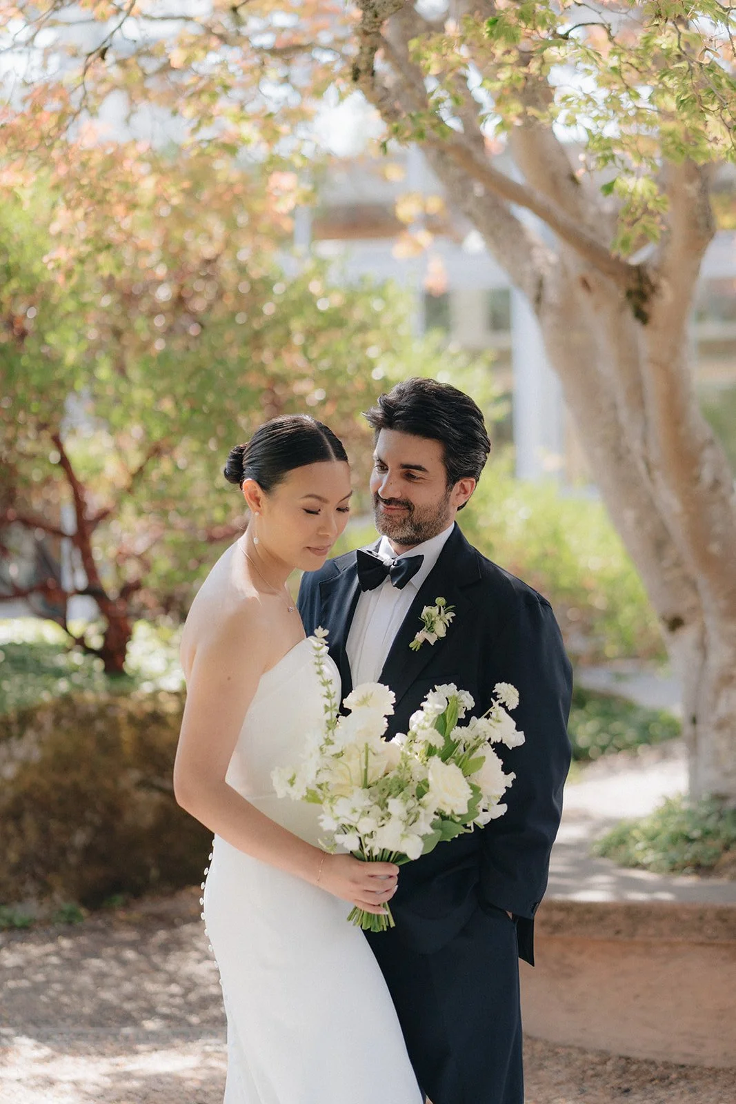 A bride and groom stand outdoors under a tree, with the bride holding a bouquet of white and green flowers, both dressed in wedding attire.
Karla makeup, Meghann hair
Photographer Kris Ladera 
Planner Lyndsay A Renee weddings
Venuew UW Center for Urb