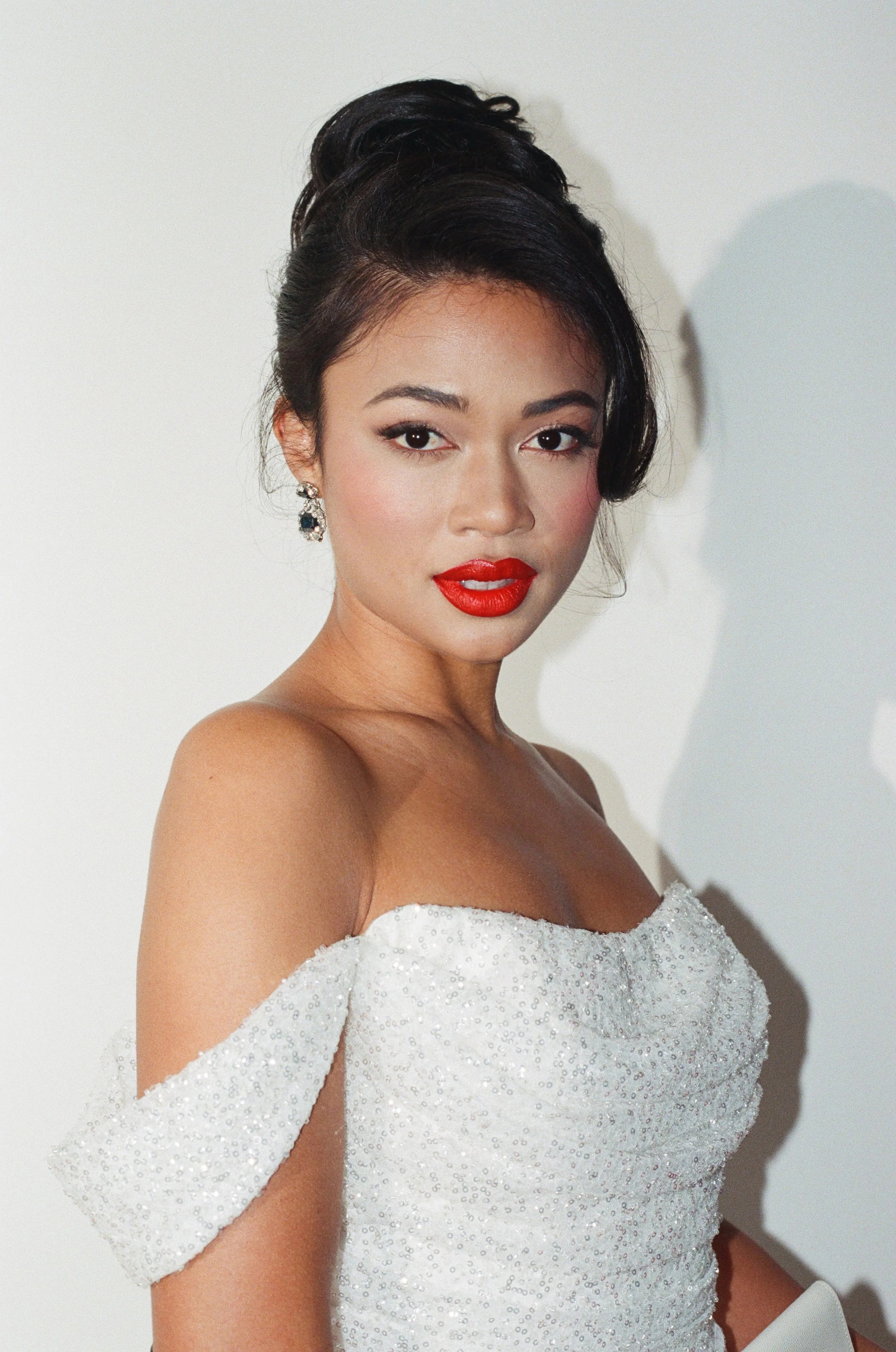 A woman with dark hair styled in an updo, wearing red lipstick, diamond earrings, and an off-shoulder white dress with sequins.