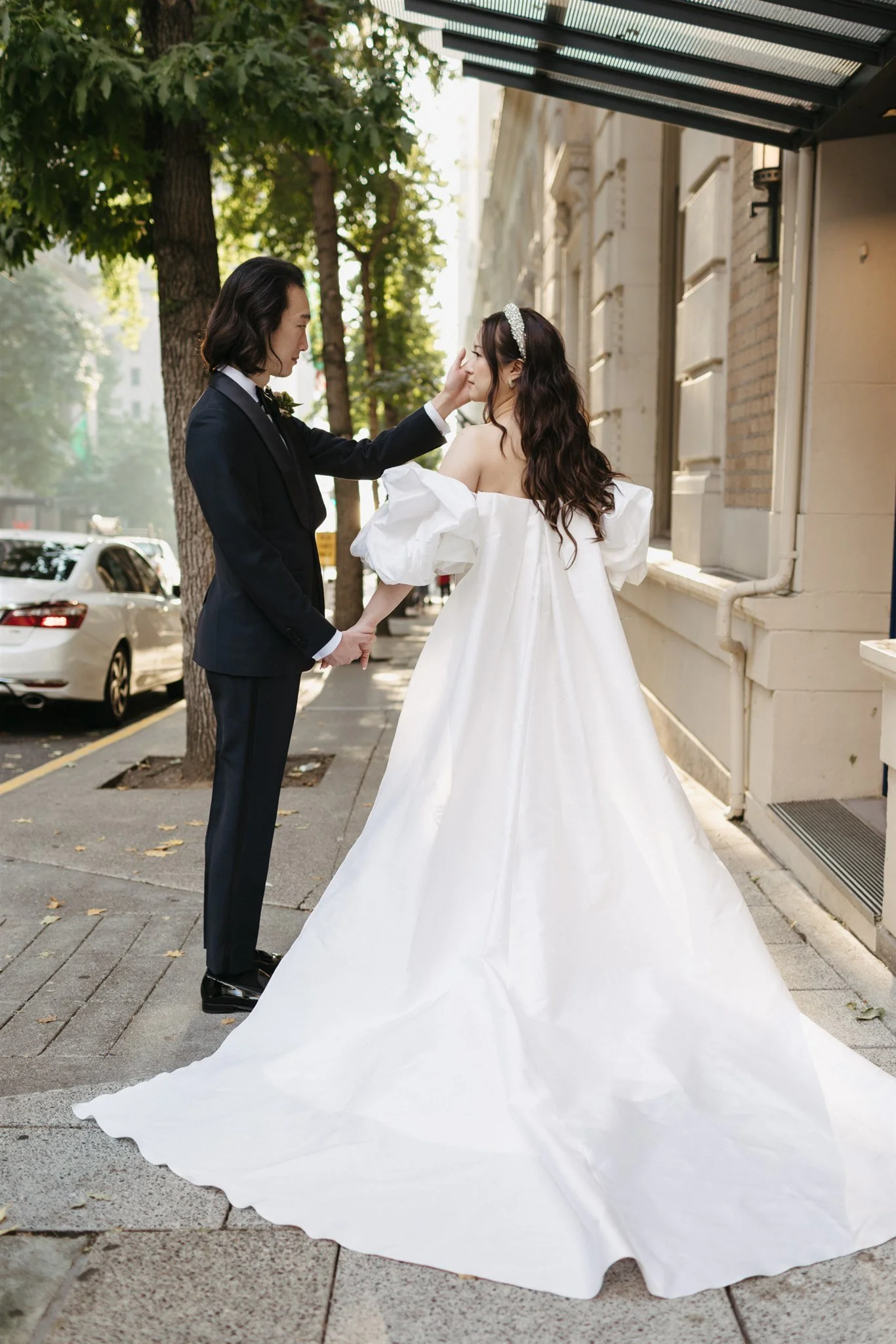 A bride and groom holding hands on a city sidewalk. The groom, dressed in a black tuxedo, gently touches the bride's face. The bride wears an off-shoulder white wedding gown with puffy sleeves and a sparkling headband. Trees and parked cars are visib