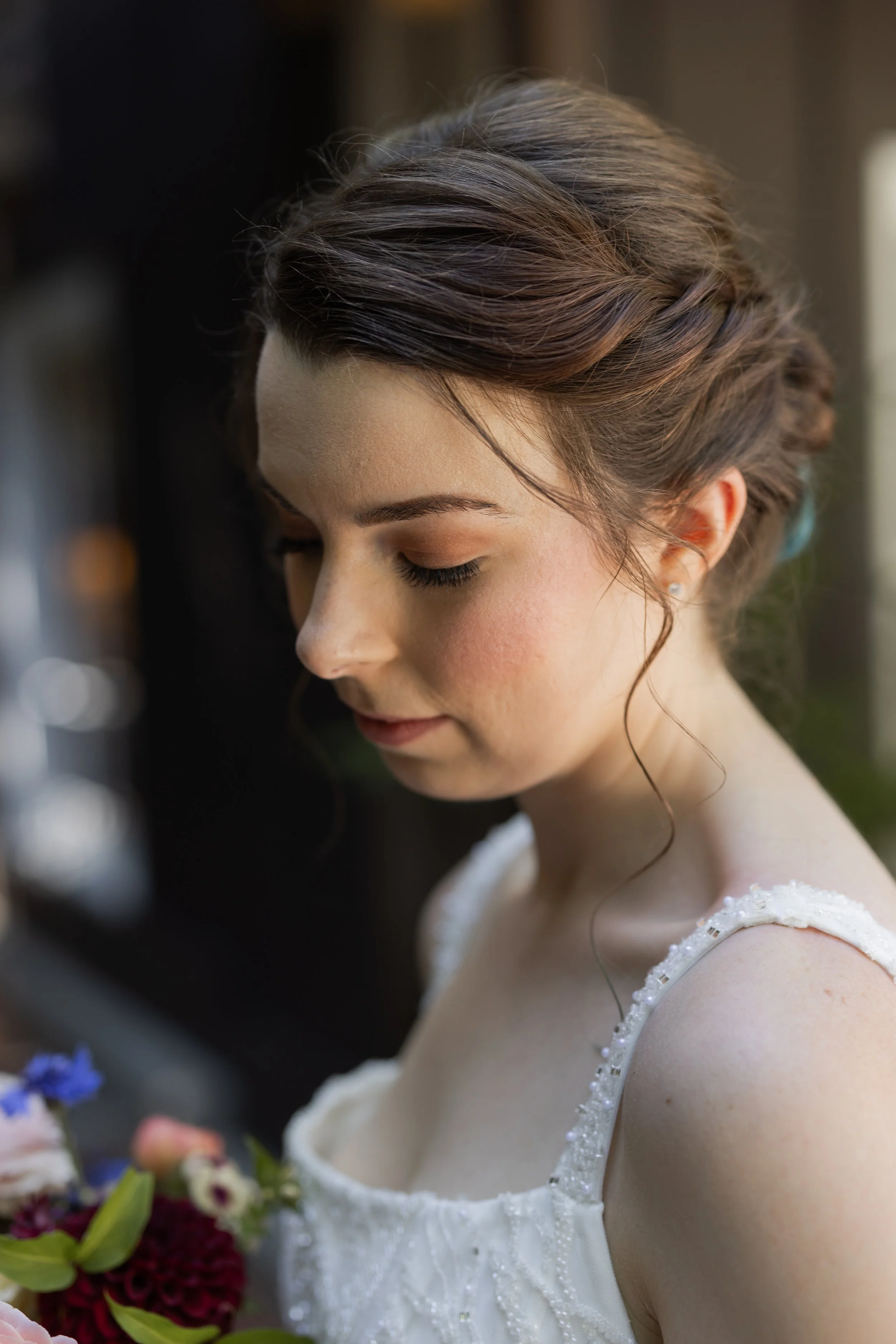 A young woman with styled brown hair and closed eyes, wearing a white dress with delicate beadwork, holding a bouquet of deep red and purple flowers.