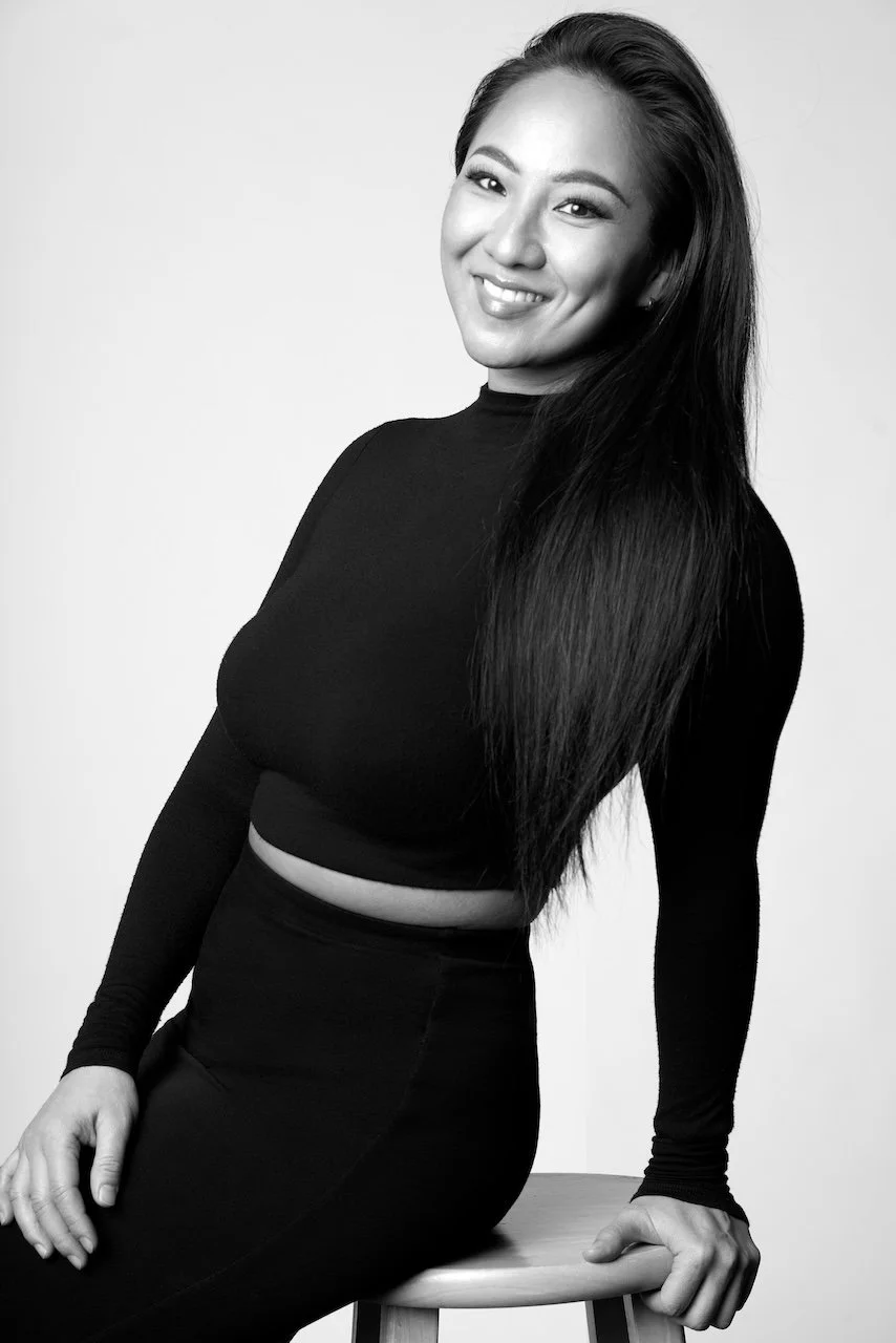 Black and white photo of a woman with long dark hair, wearing a fitted long-sleeve top and high-waisted pants, sitting on a stool and smiling at the camera.