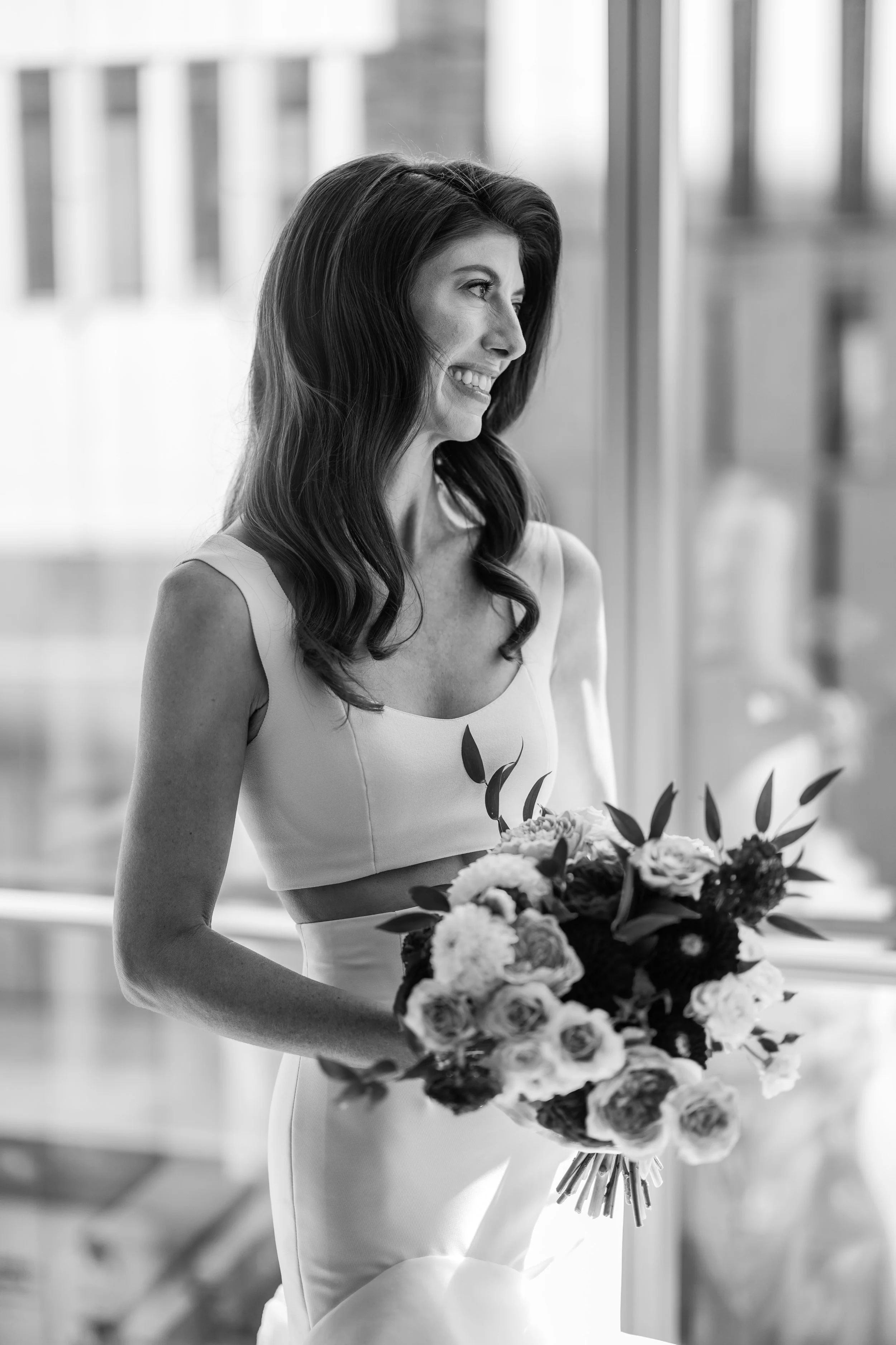 A smiling woman with long wavy hair, wearing a sleeveless dress, holding a large bouquet of flowers, standing indoors near a window.