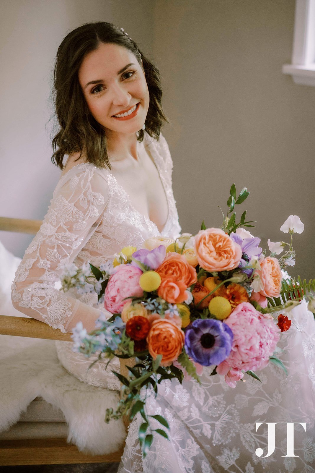 A woman in a white lace wedding dress smiling while holding a colorful bouquet of flowers, sitting on a bench with a white furry throw.