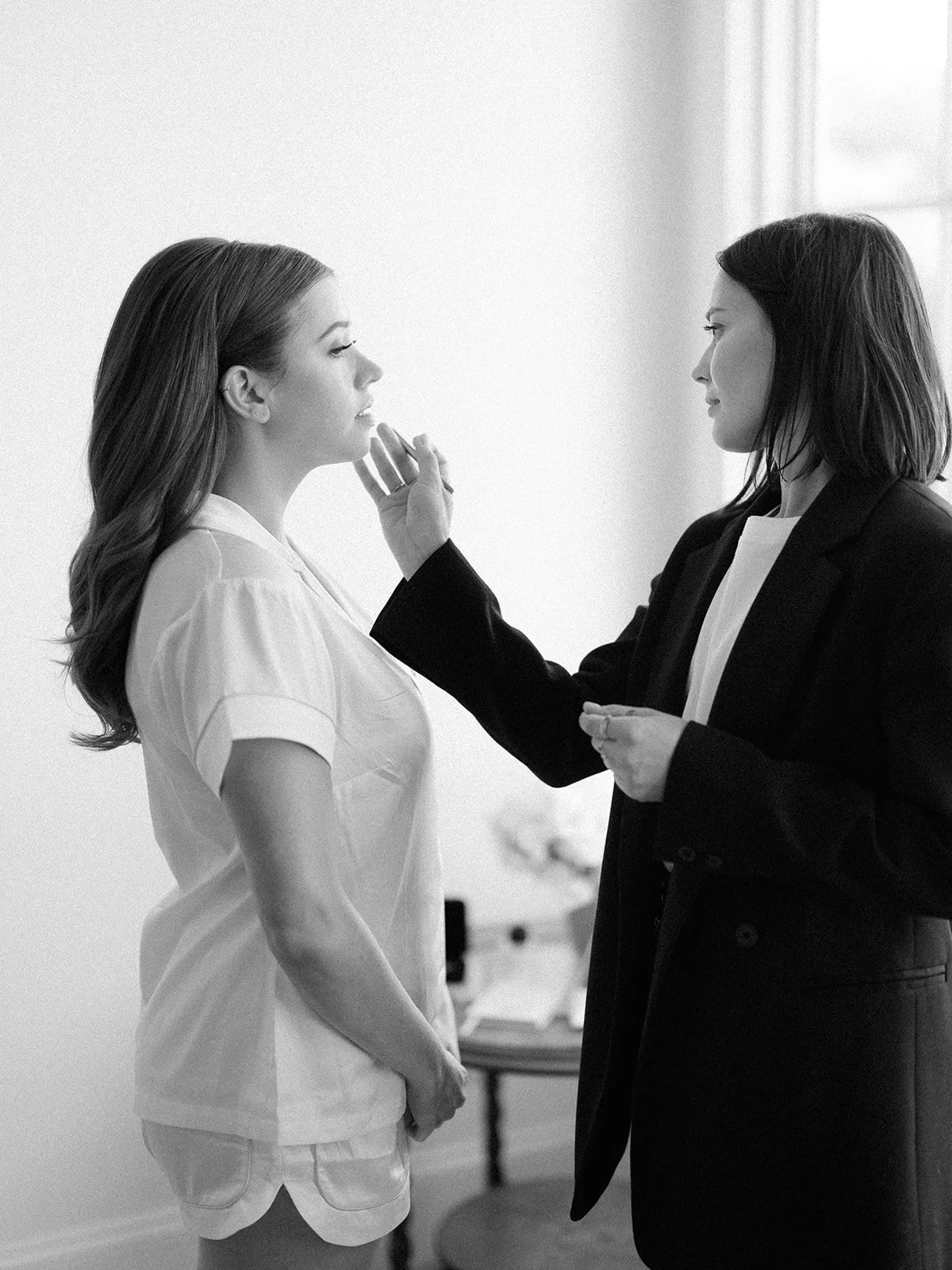 A woman in a white uniform is having her makeup or face checked by another woman in a dark suit, in a bright room with natural light.