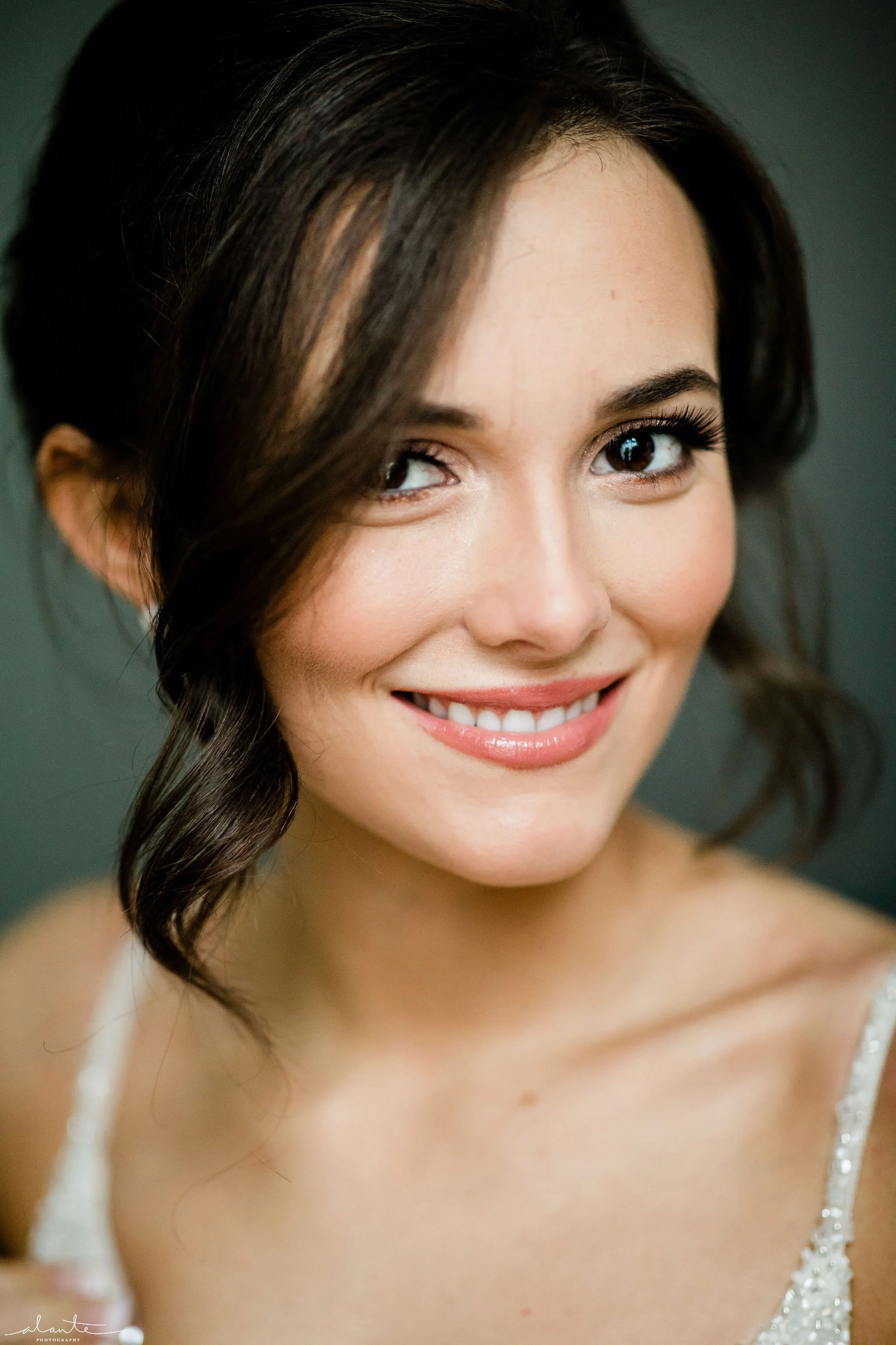 Close-up of a smiling woman with dark hair styled in loose curls, wearing a white dress with beaded straps, against a dark background.