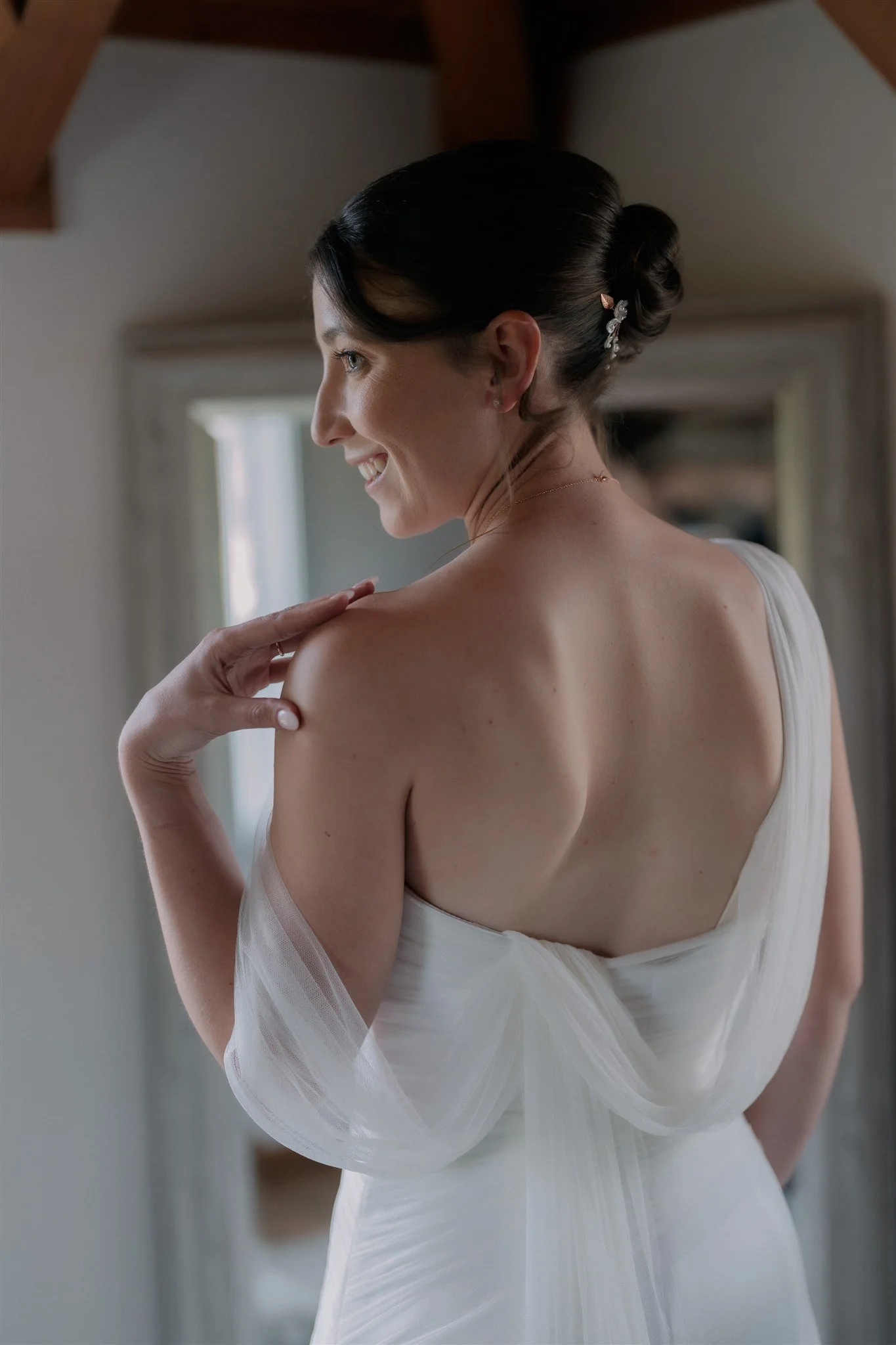 Close-up of a bride with dark hair styled in an elegant bun, wearing a white dress with sheer sleeves, smiling softly as she touches her shoulder.