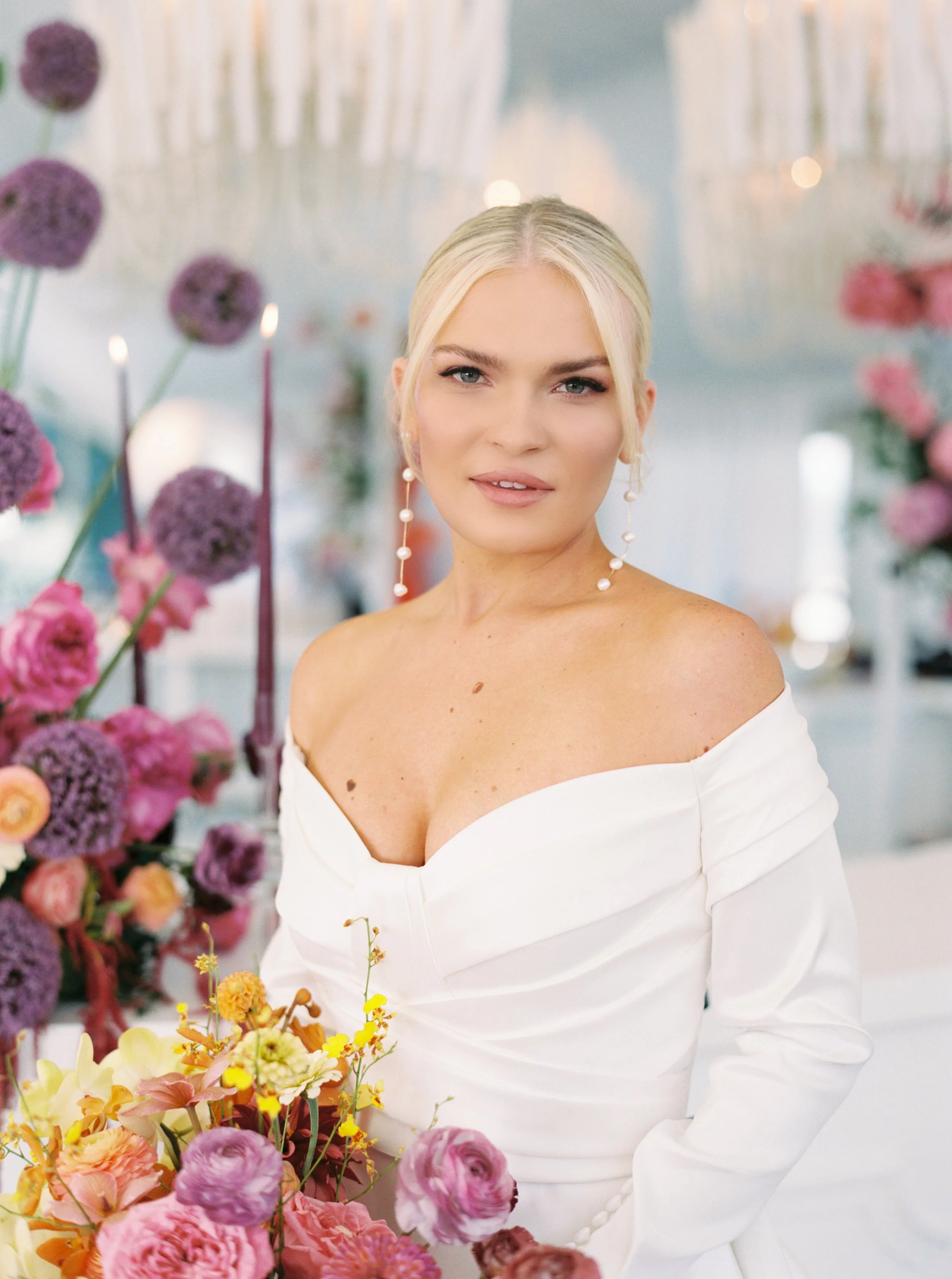 A woman in a white off-shoulder dress with long earrings stands amidst a display of colorful flowers, including pink, purple, and yellow blossoms, with a blurred background of candles and flowers.