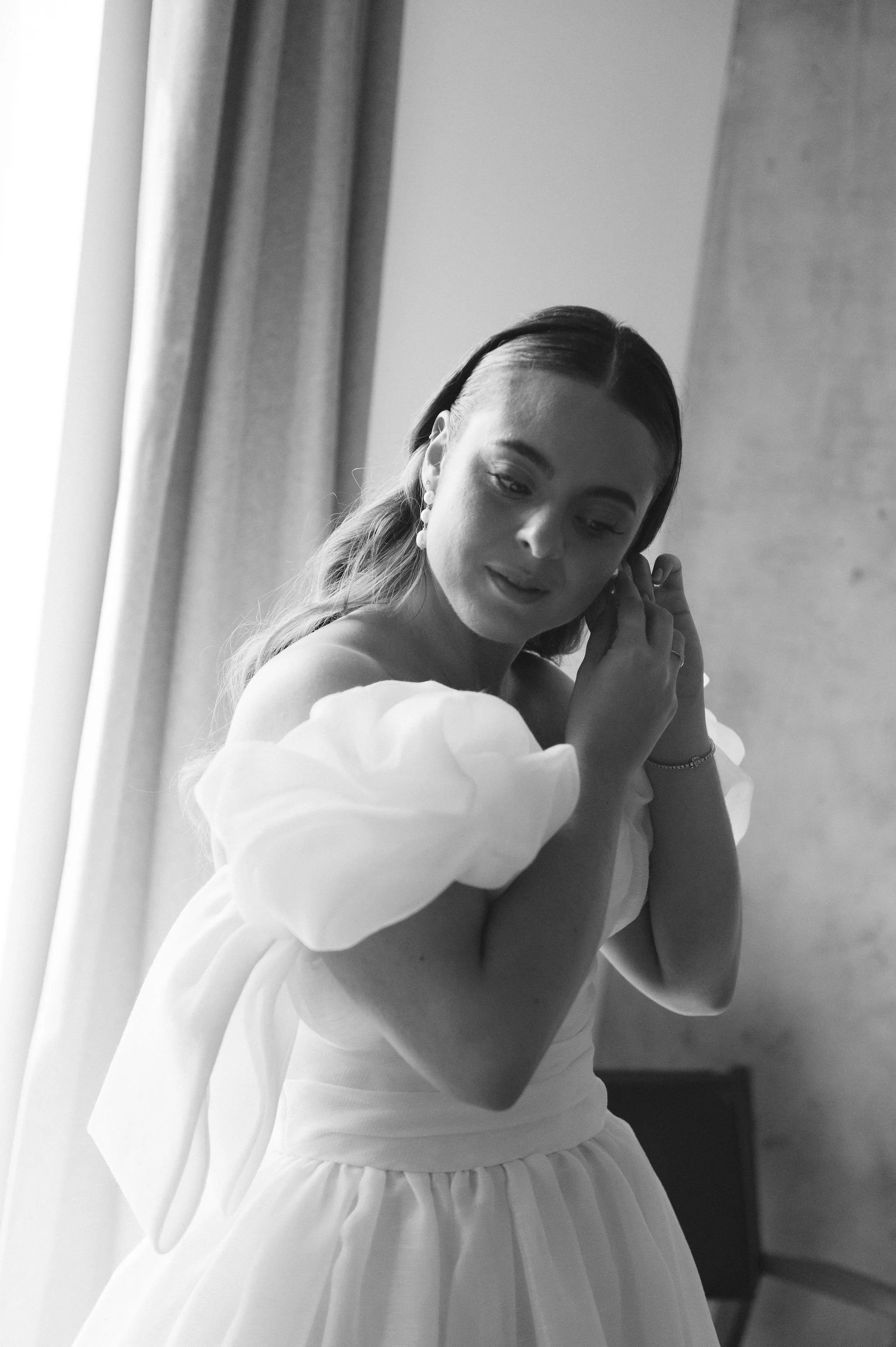 A black-and-white photo of a woman with long hair, wearing earrings, a ruffled off-shoulder top, and a skirt, touching her ear and looking down, standing near a window with curtains.
Makeup Katya
Hair Meghhann
Venue Admiral's house
Planner Amber Soot