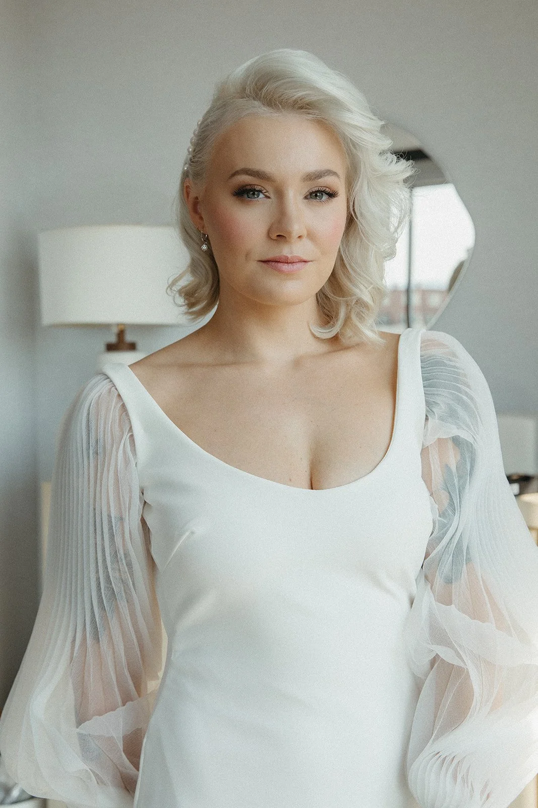 A woman with blonde, wavy hair and blue eyes, wearing a white dress with sheer, pleated sleeves, standing indoors in a well-lit room with a lamp and window in the background.