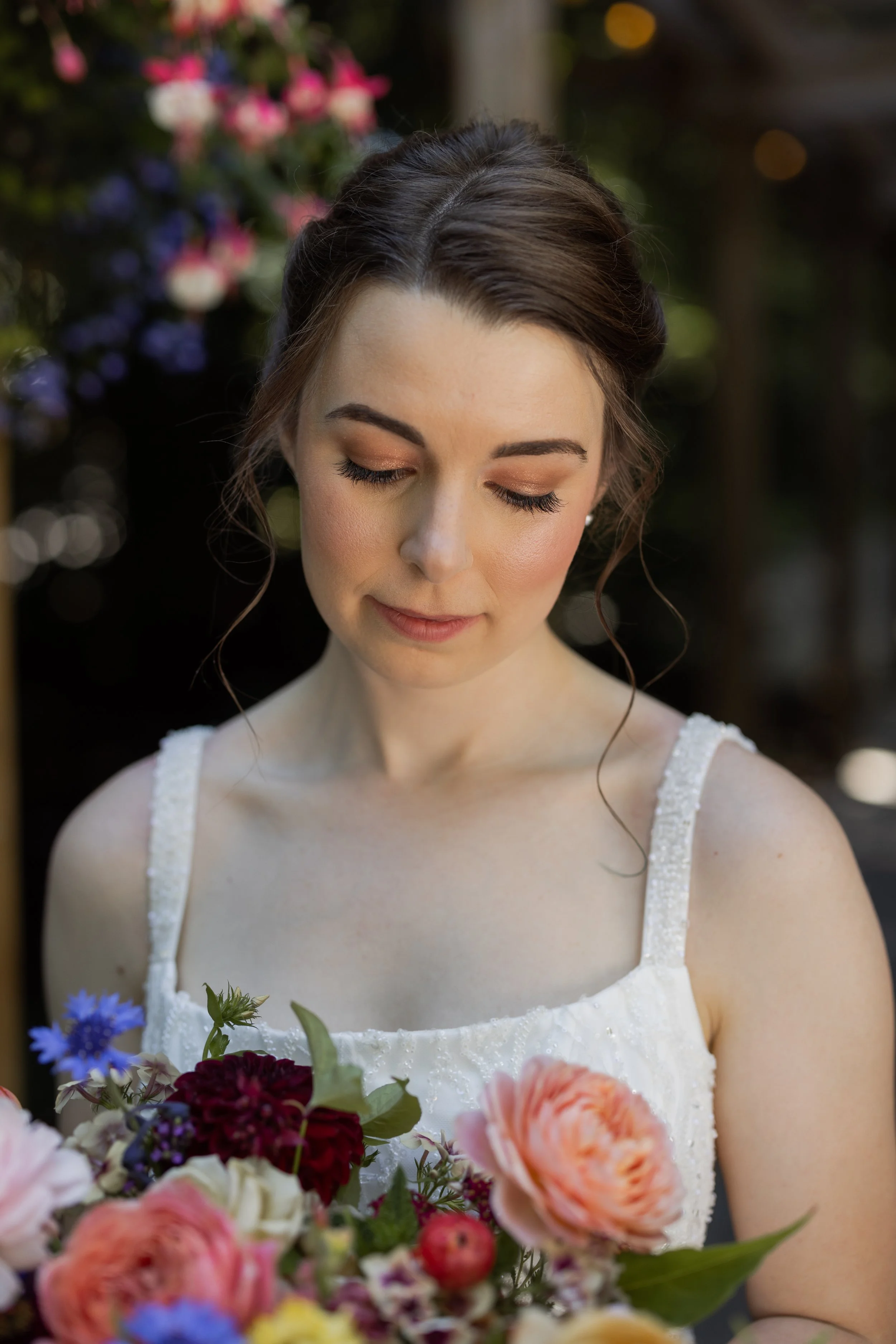 A woman in a white dress with beaded straps is holding a large bouquet of colorful flowers, with her eyes closed and a gentle smile.