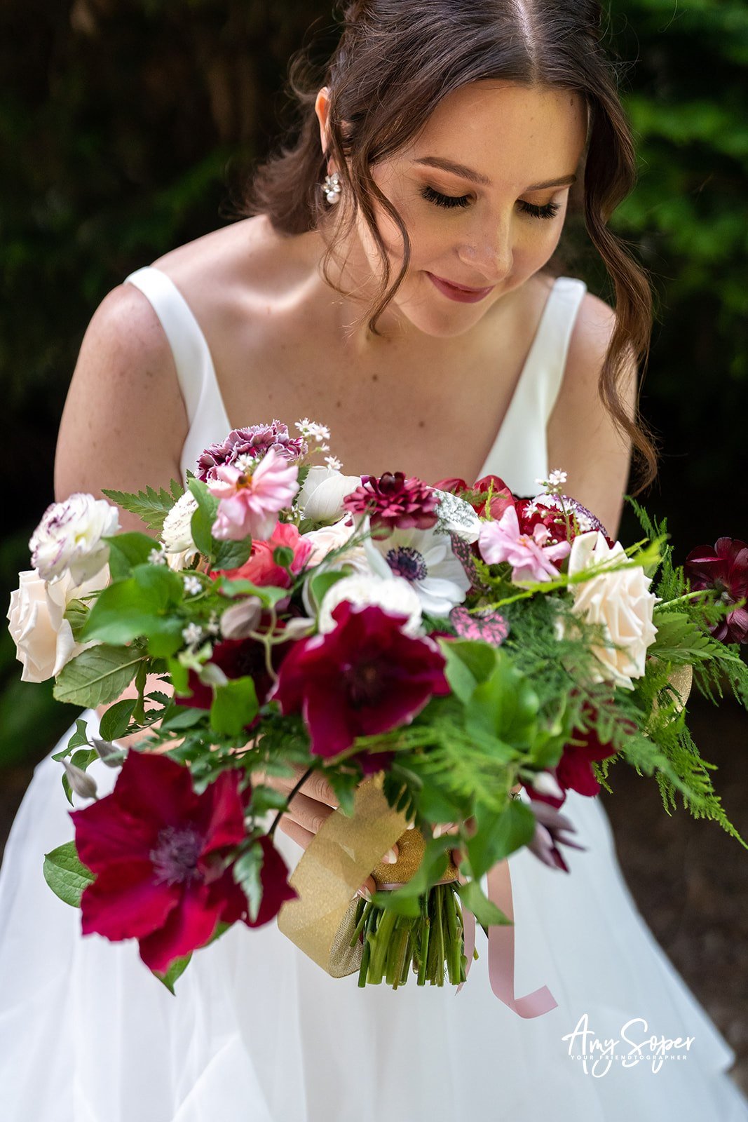 A bride in a white dress holding a large bouquet of pink, white, and red flowers with green foliage, standing outdoors with blurred greenery in the background.