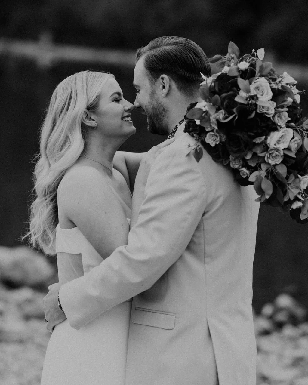 A black-and-white photograph of a bride and groom in a close embrace, smiling at each other, with a large bouquet of flowers on the groom's shoulder.