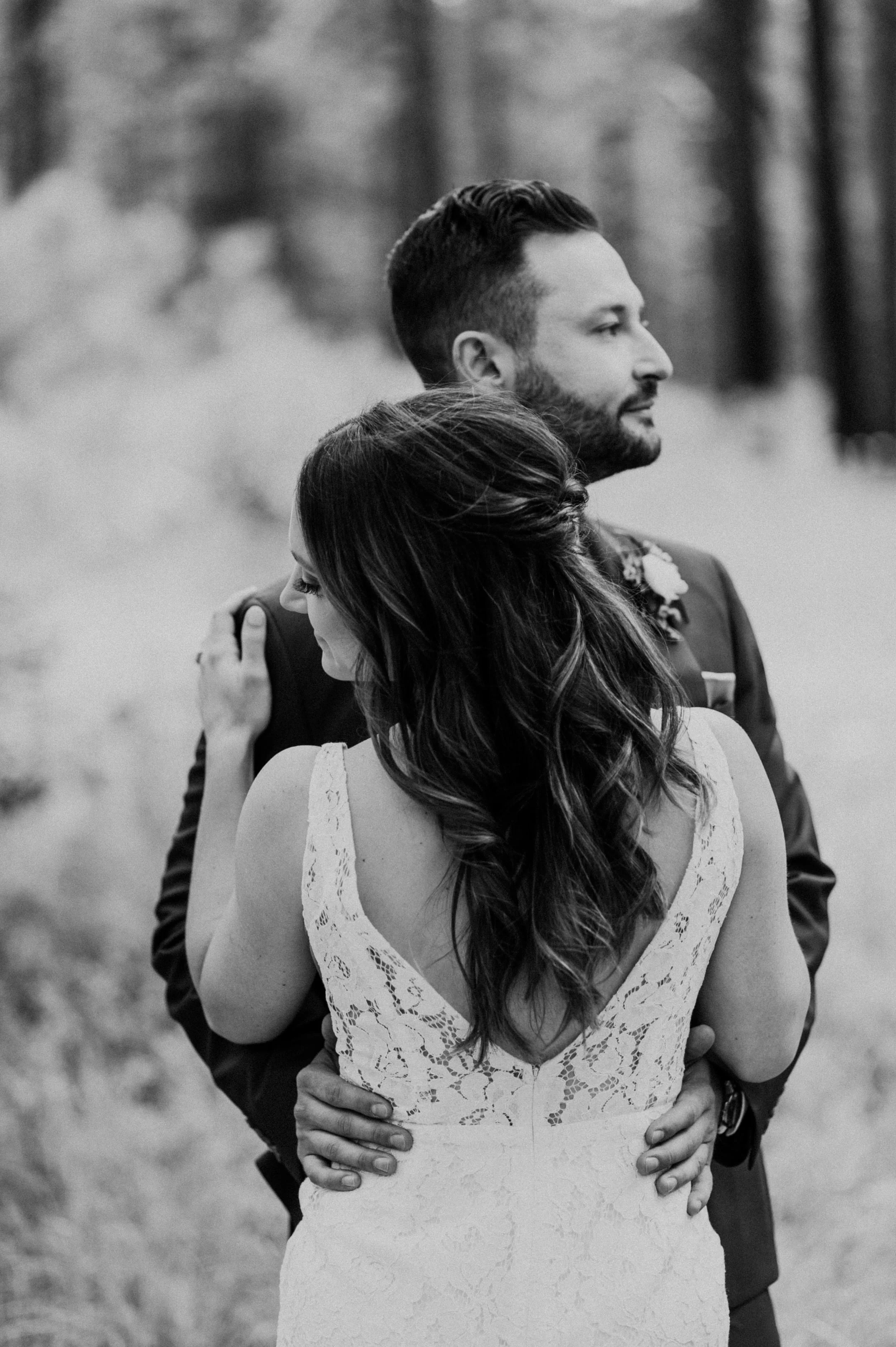 A man and woman are embracing outdoors. The woman has long, wavy hair and is wearing a lacy, sleeveless dress. The man has short, dark hair and a beard, and is wearing a suit. They are holding each other closely, with the man's hands on the woman’s w