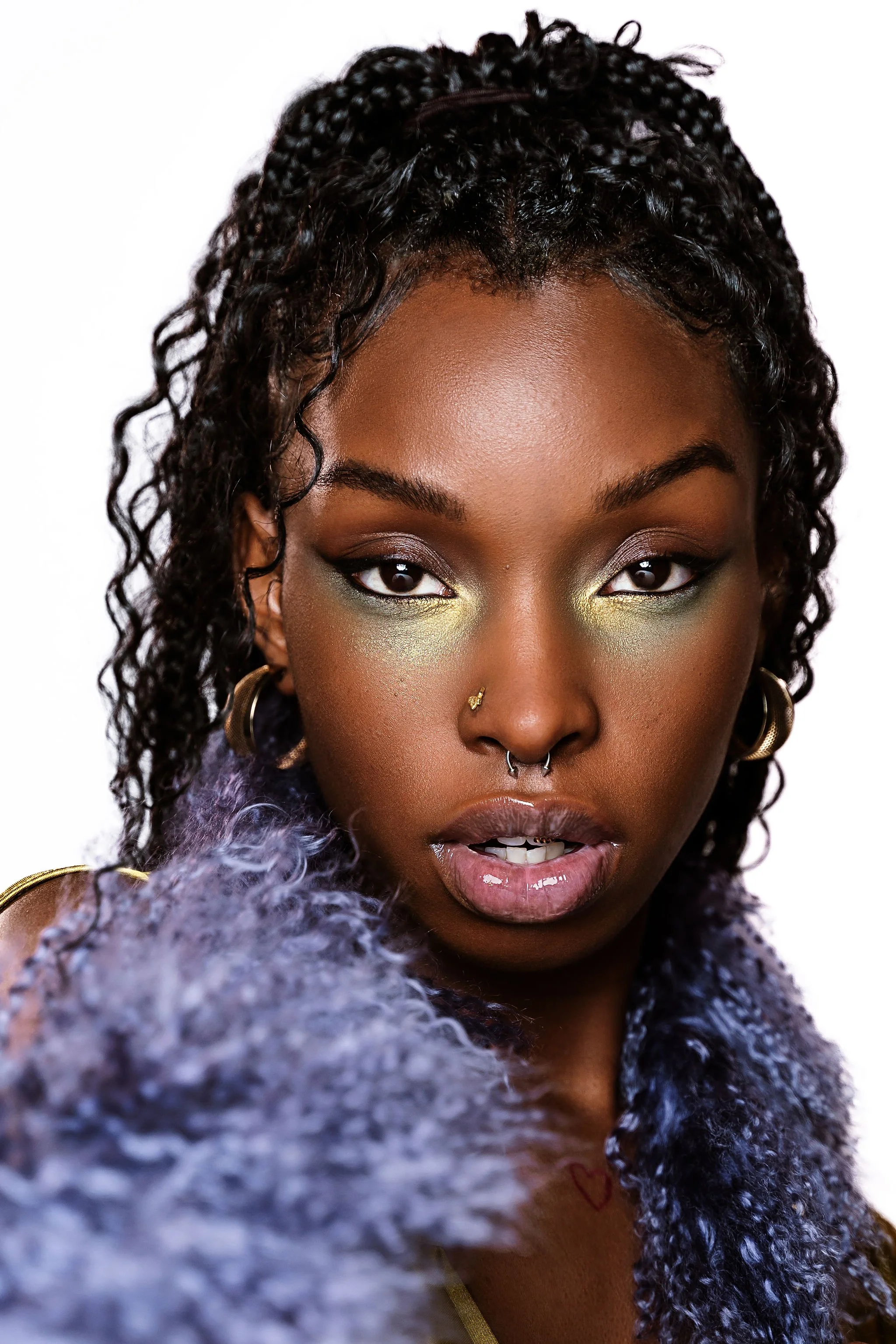 A close-up portrait of a woman with dark curly hair styled in a high ponytail, wearing colorful eye makeup with green and gold eyeshadow, hoop earrings, and a textured purple fur garment.